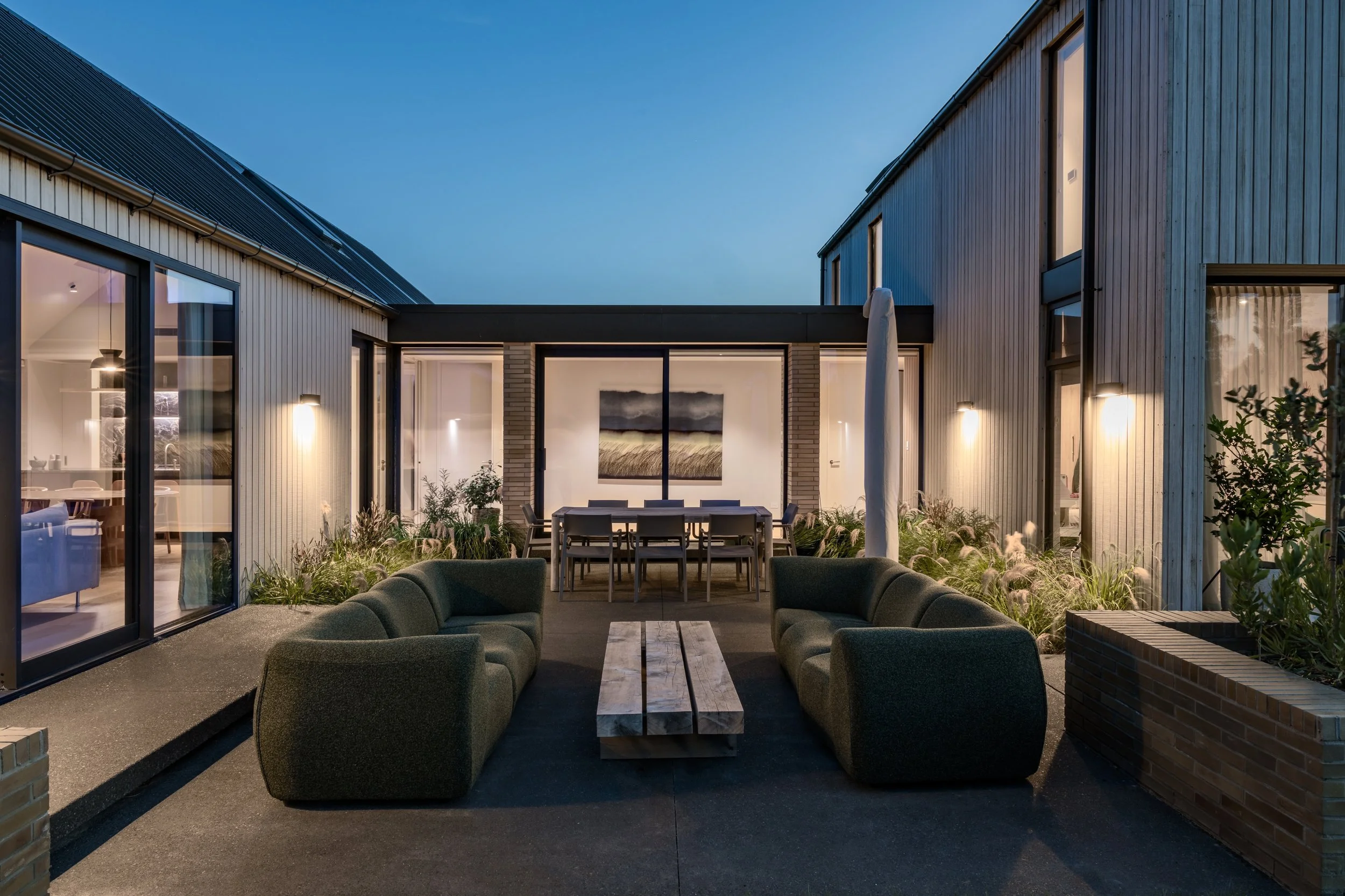 Modern outdoor patio area at dusk with curved seating, a wooden table, and an open view of two contemporary houses with wood siding and large windows.