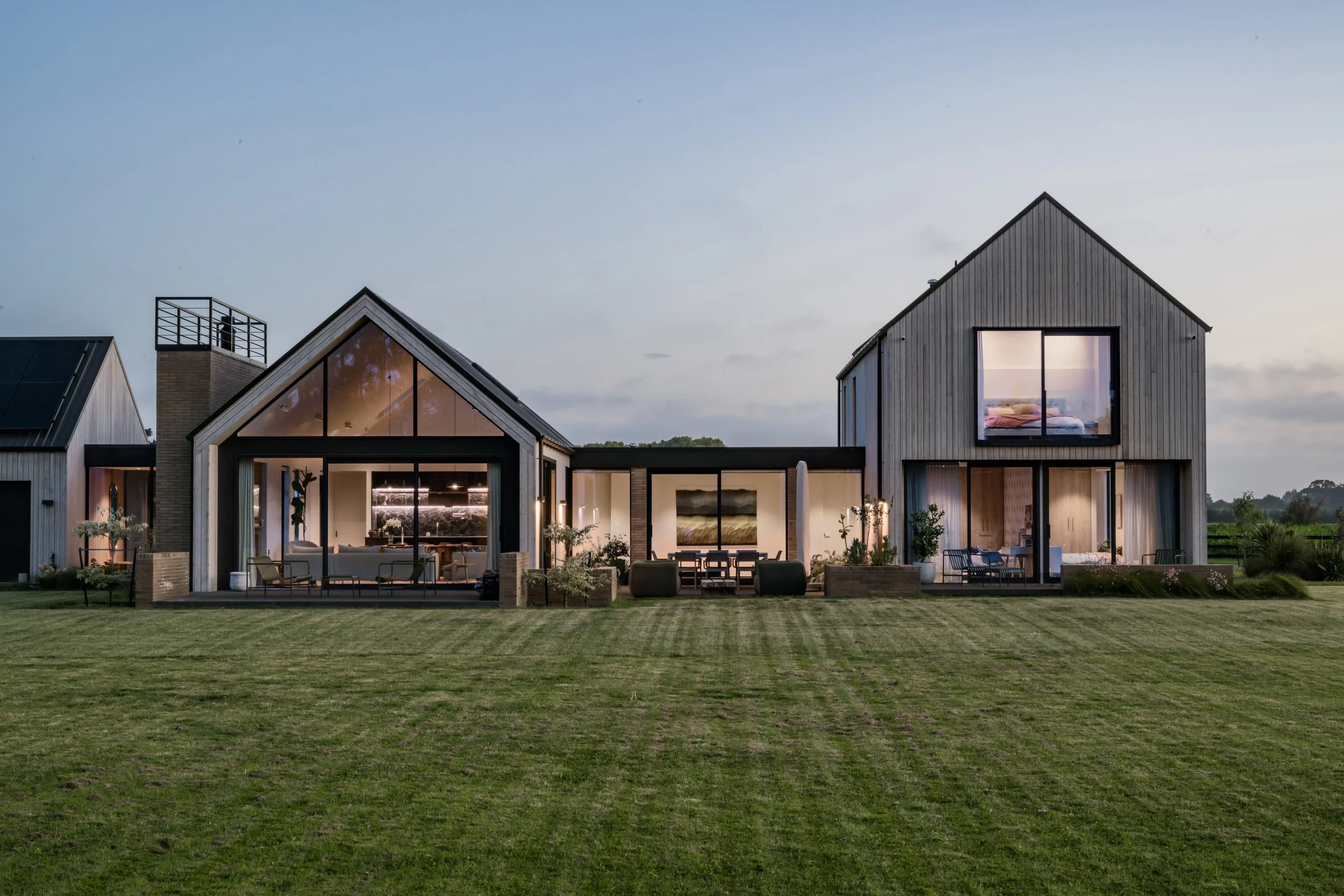 Modern house with large glass windows and wooden exterior, illuminated inside, surrounded by a well-maintained lawn, during dusk.