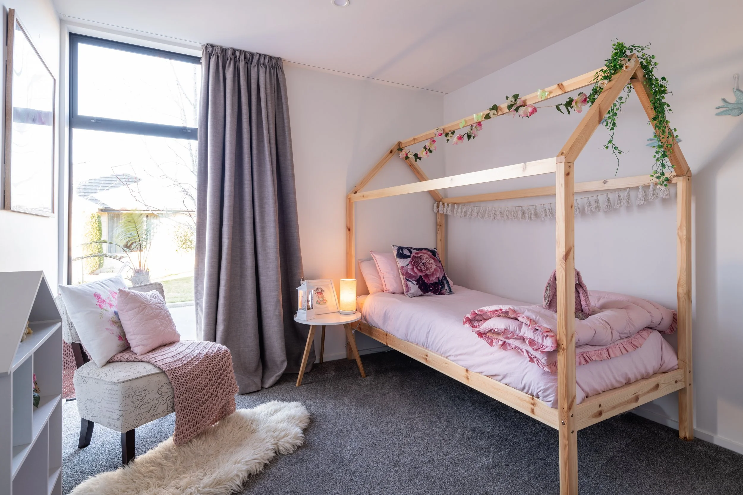 A cozy bedroom with a house-shaped bed frame decorated with pink flowers and greenery, pink bedding, a small side table with a lamp, a white armchair with pink pillows and a knitted throw, a soft area rug, and a large window with gray curtains.