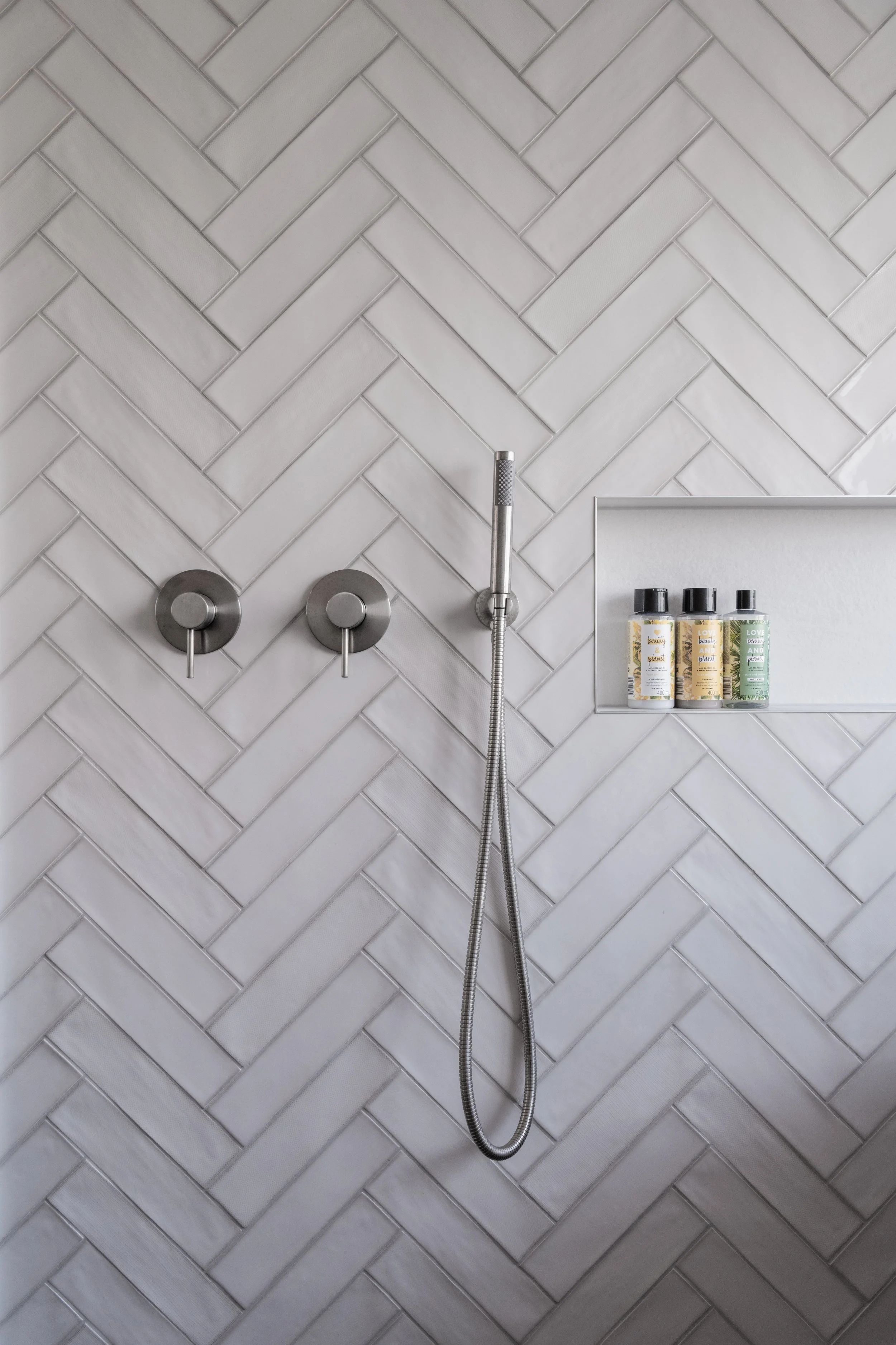 Shower wall with white herringbone tiles, a handheld showerhead, two silver controls, and a built-in shelf with three bottles of bath products.