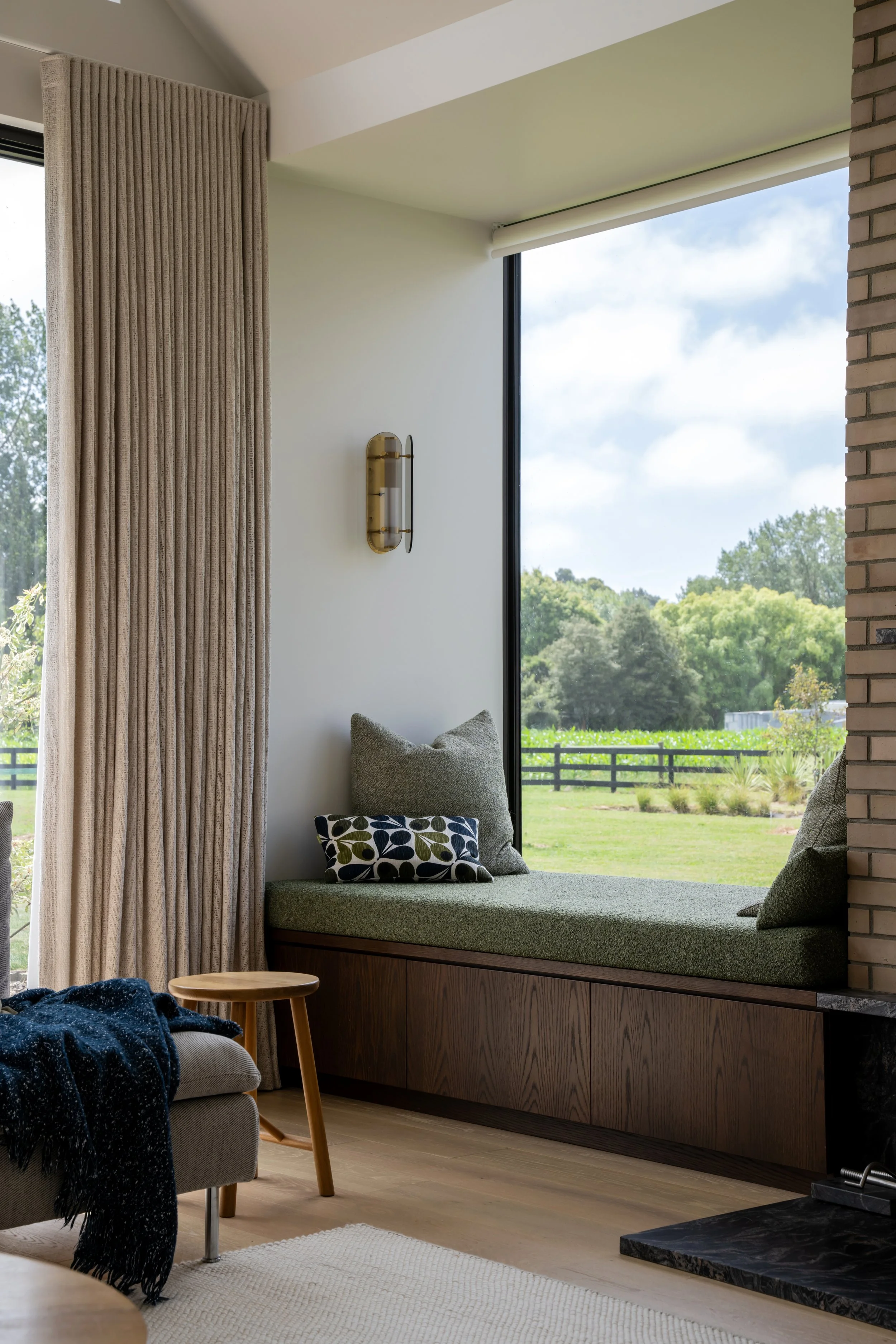 Living room with large window showcasing green trees and sky, beige curtains, a built-in window seat with cushions, a small wooden stool, a gray armchair with a dark blue blanket, and a black marble fireplace.