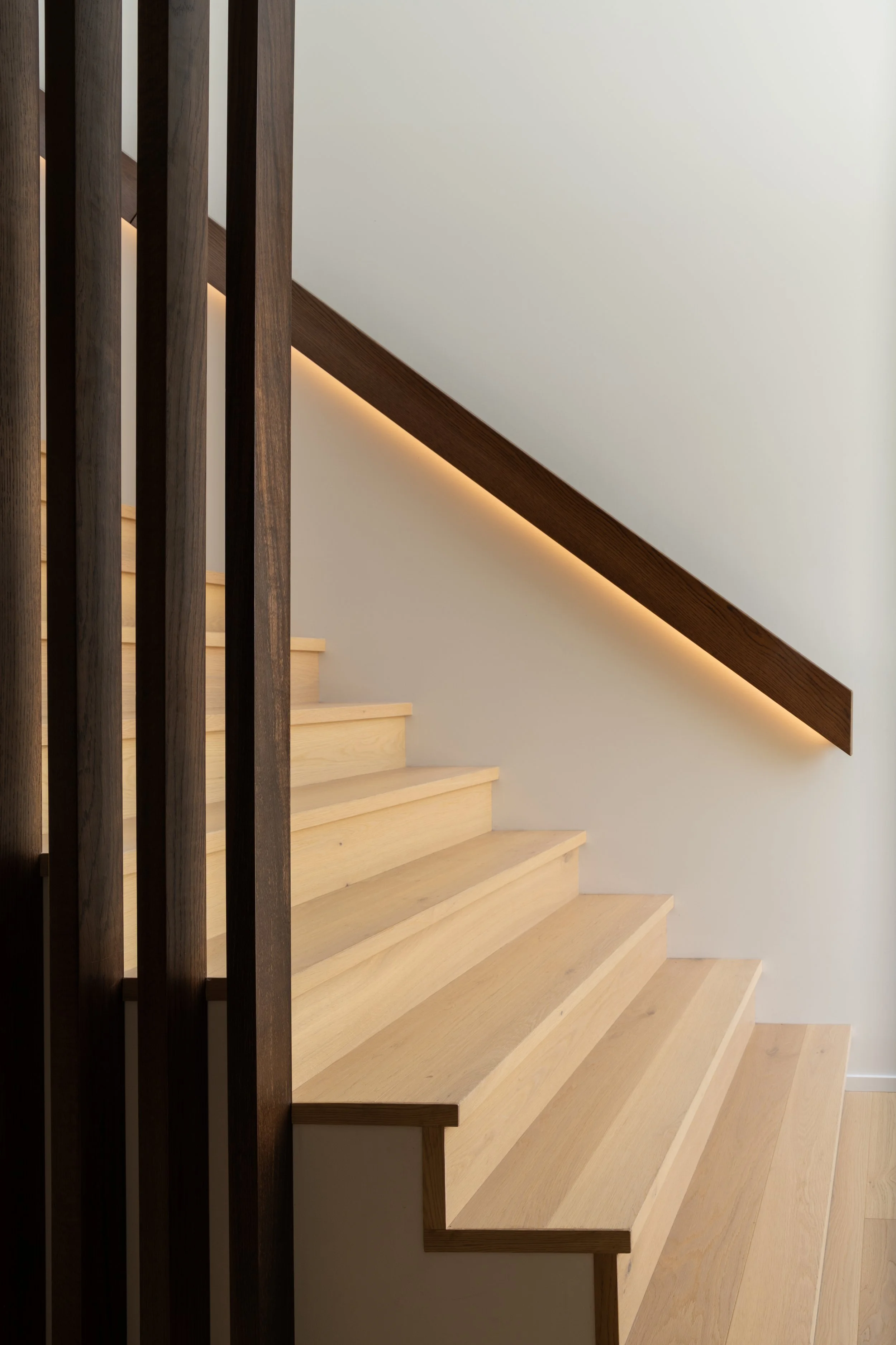 Wooden staircase with light-colored steps and a dark wooden handrail against a white wall, with integrated lighting underneath the handrail.