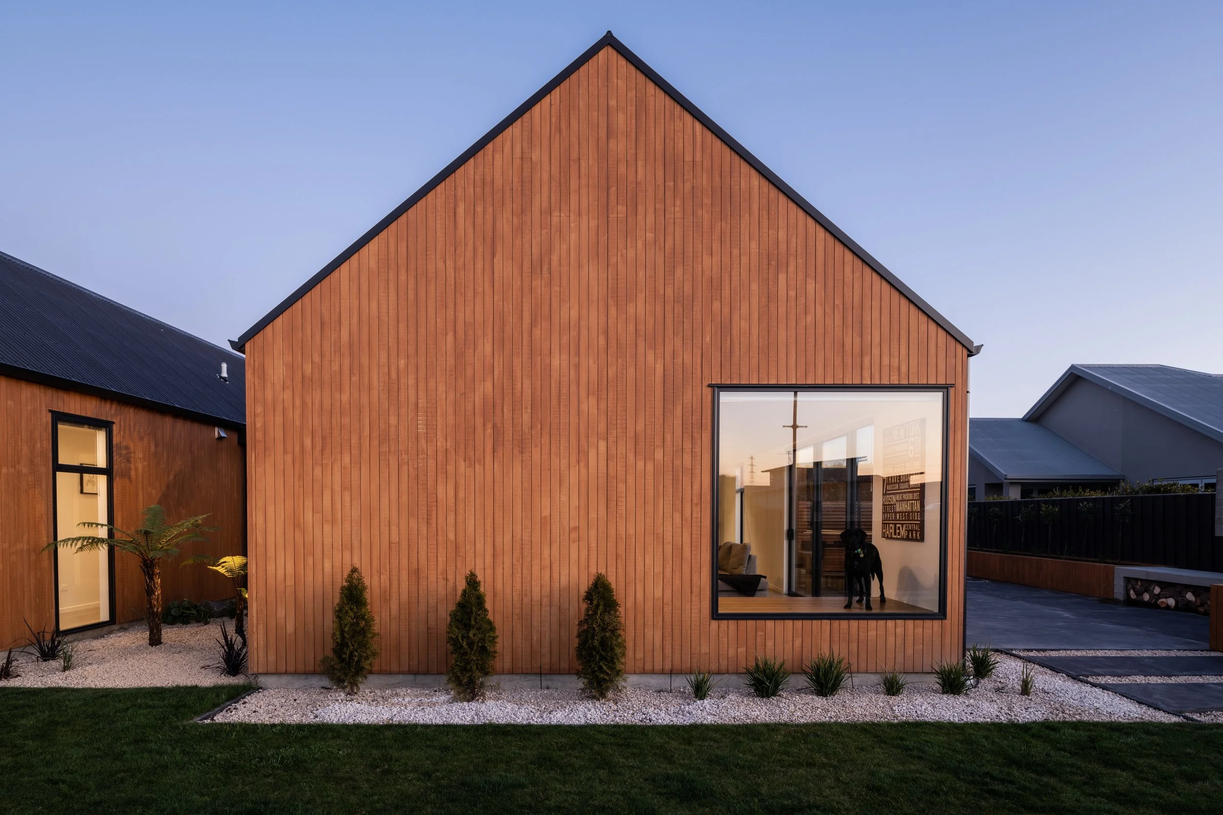 Modern house with a wooden exterior, large front window, small plants, and a well-maintained lawn in the evening.