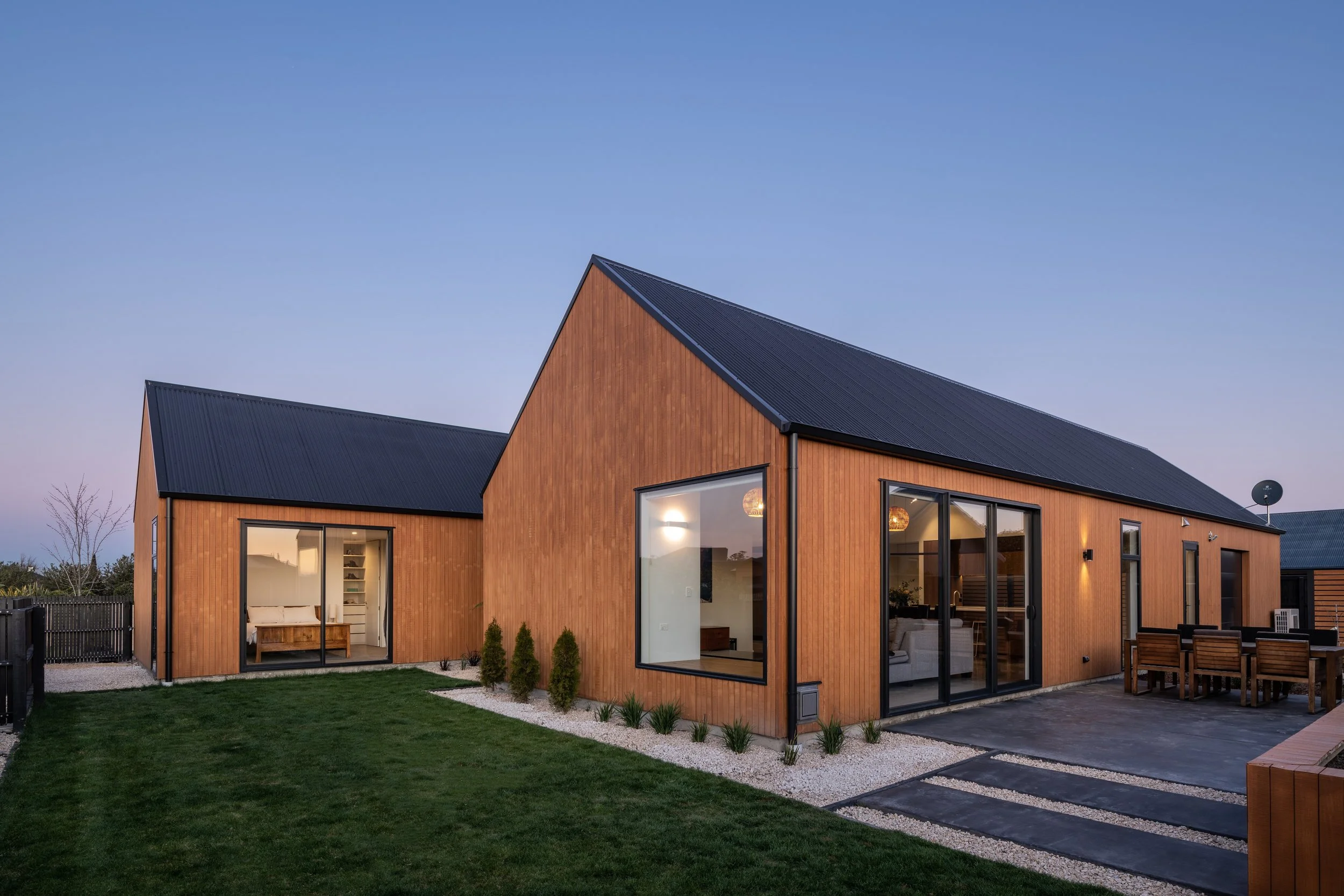Modern house with wooden exterior, large glass windows, outdoor dining area, well-maintained lawn, and a clear sky at dusk.