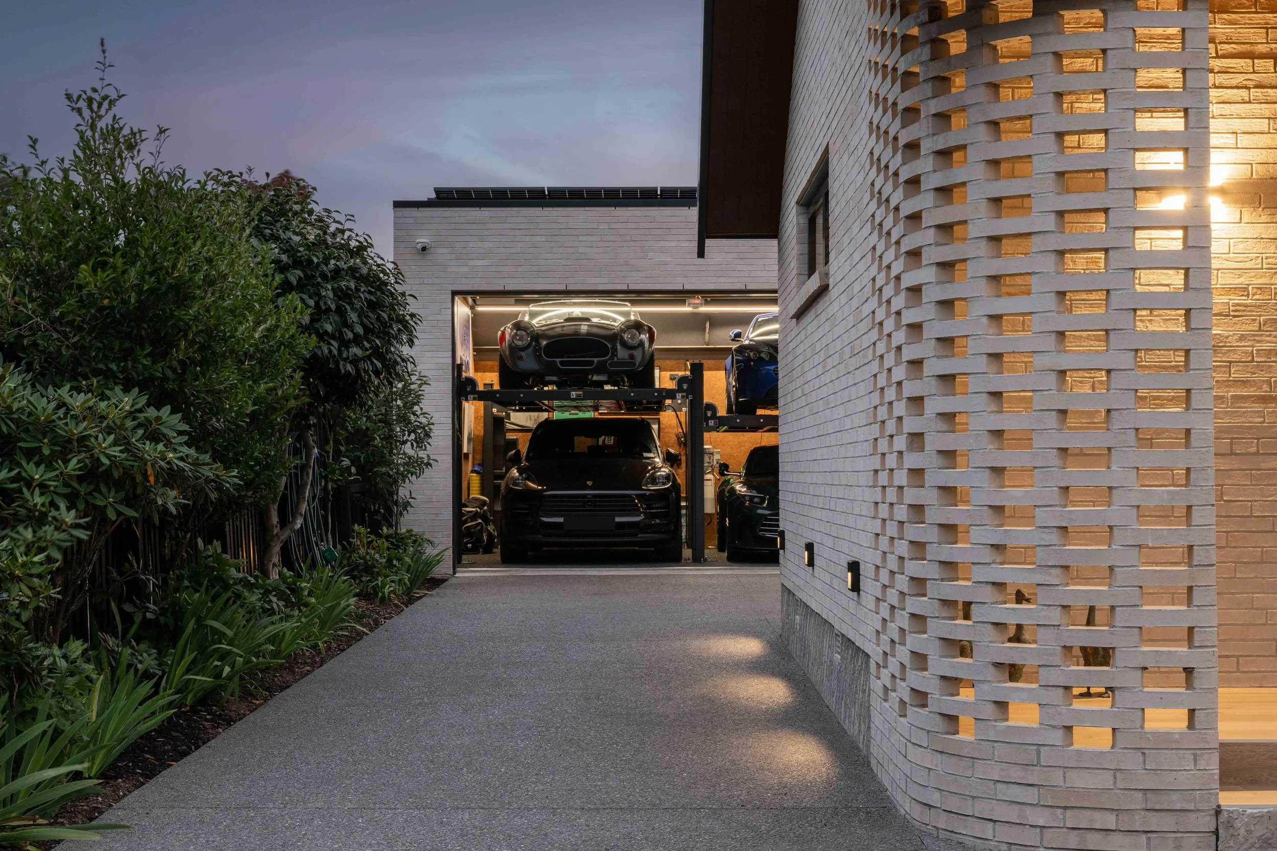 Modern house garage with a sports car on a lift and a sleek brick and illuminated wall on the right side, surrounded by greenery and an overcast sky.