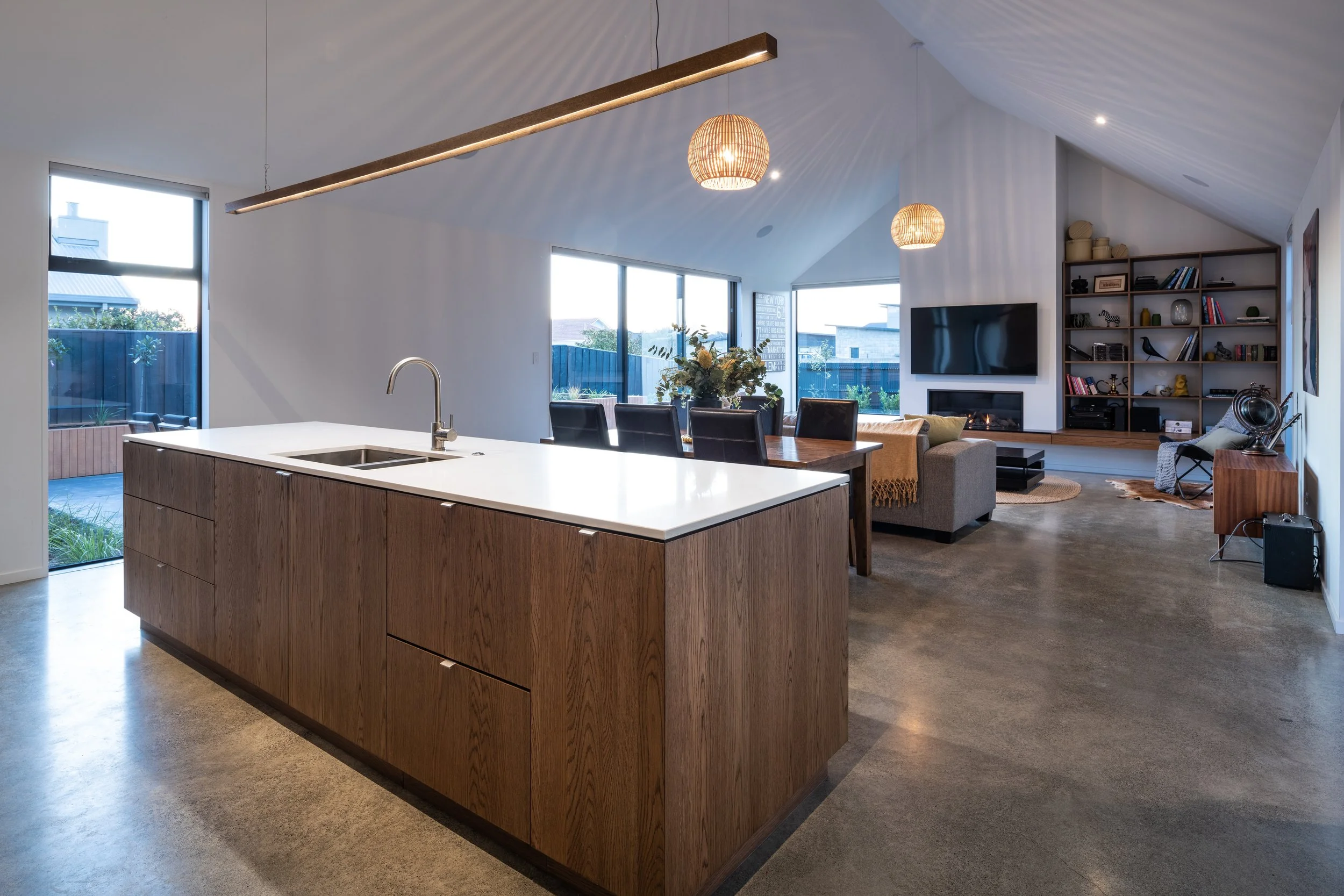 Open-concept living space with a modern kitchen island, dining area, and living room with a fireplace and bookshelf, large windows, and minimalist decor.