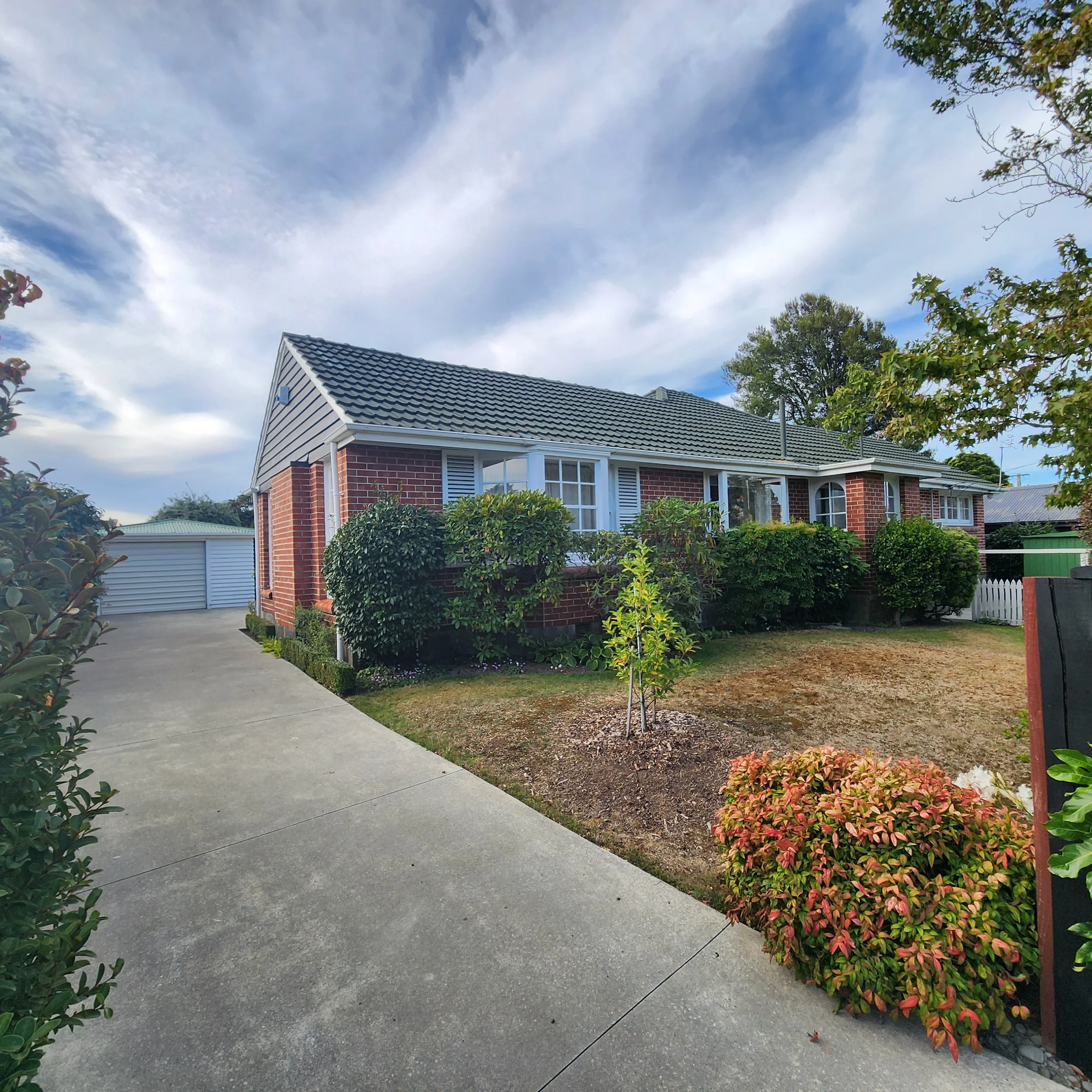 A suburban residential house with a tiled roof, surrounded by a garden with bushes and small trees, under a sky with scattered clouds.
