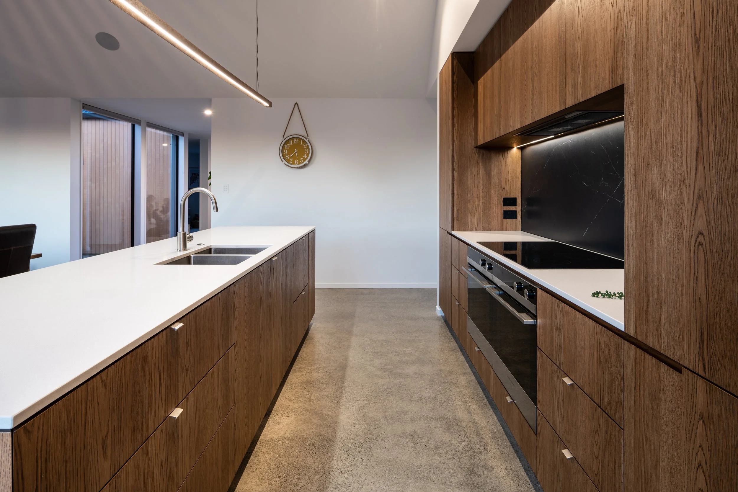 Modern kitchen with wooden cabinets, white countertops, black flat stovetop, built-in oven, and stainless steel sink, with sliding glass doors and a wall clock in the background.