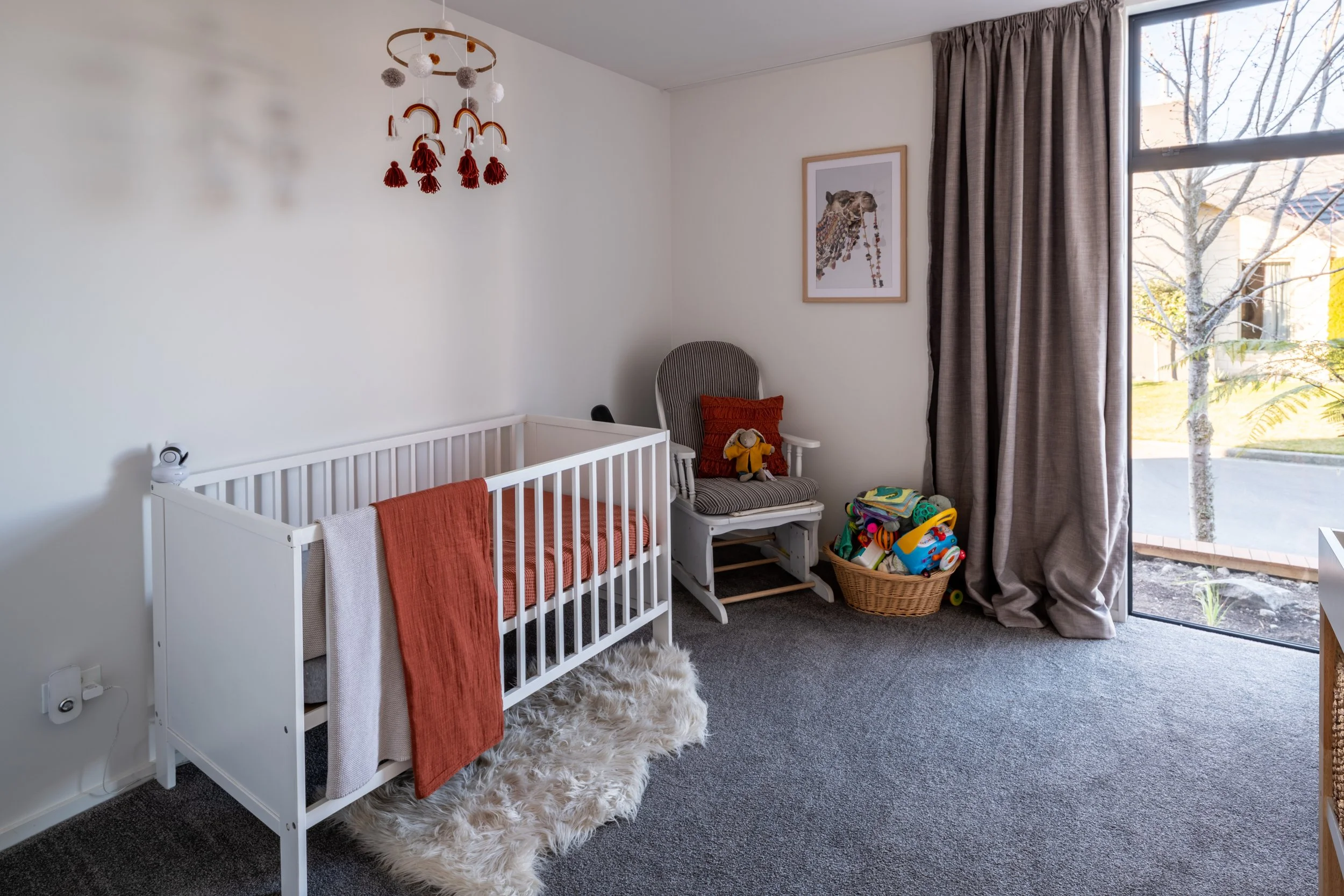 Nursery with white crib, rocking chair, basket of toys, and large window with beige curtains.