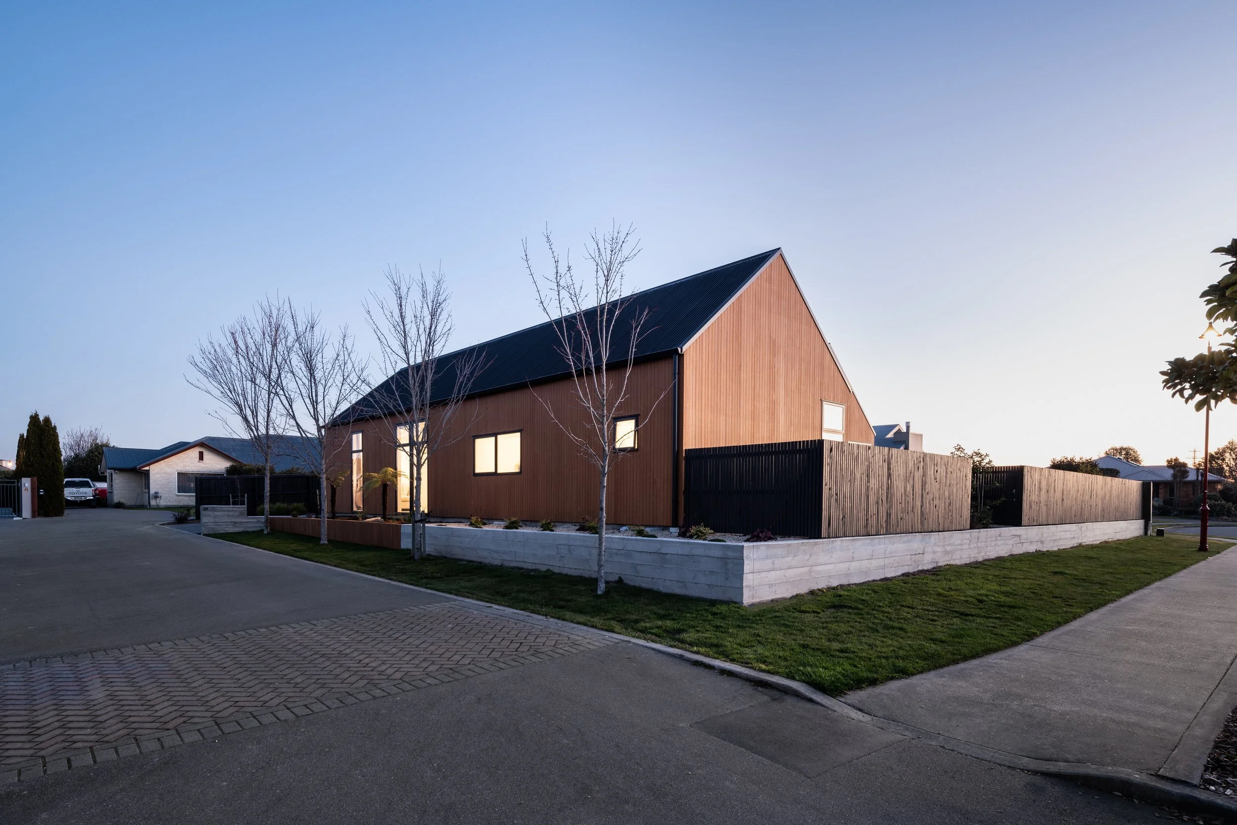 Modern house with wooden exterior, sloped black roof, and front yard with trees and green lawn, at dusk.