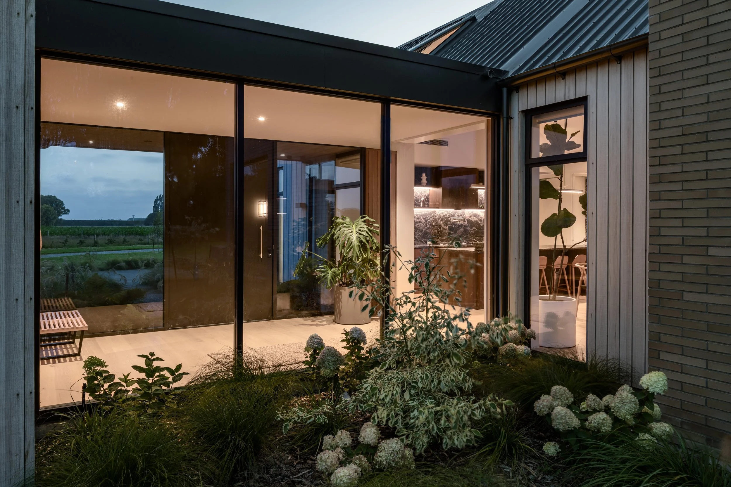 Modern house exterior with large glass windows, visible interior lighting, and a small garden with various plants and flowers at dusk.