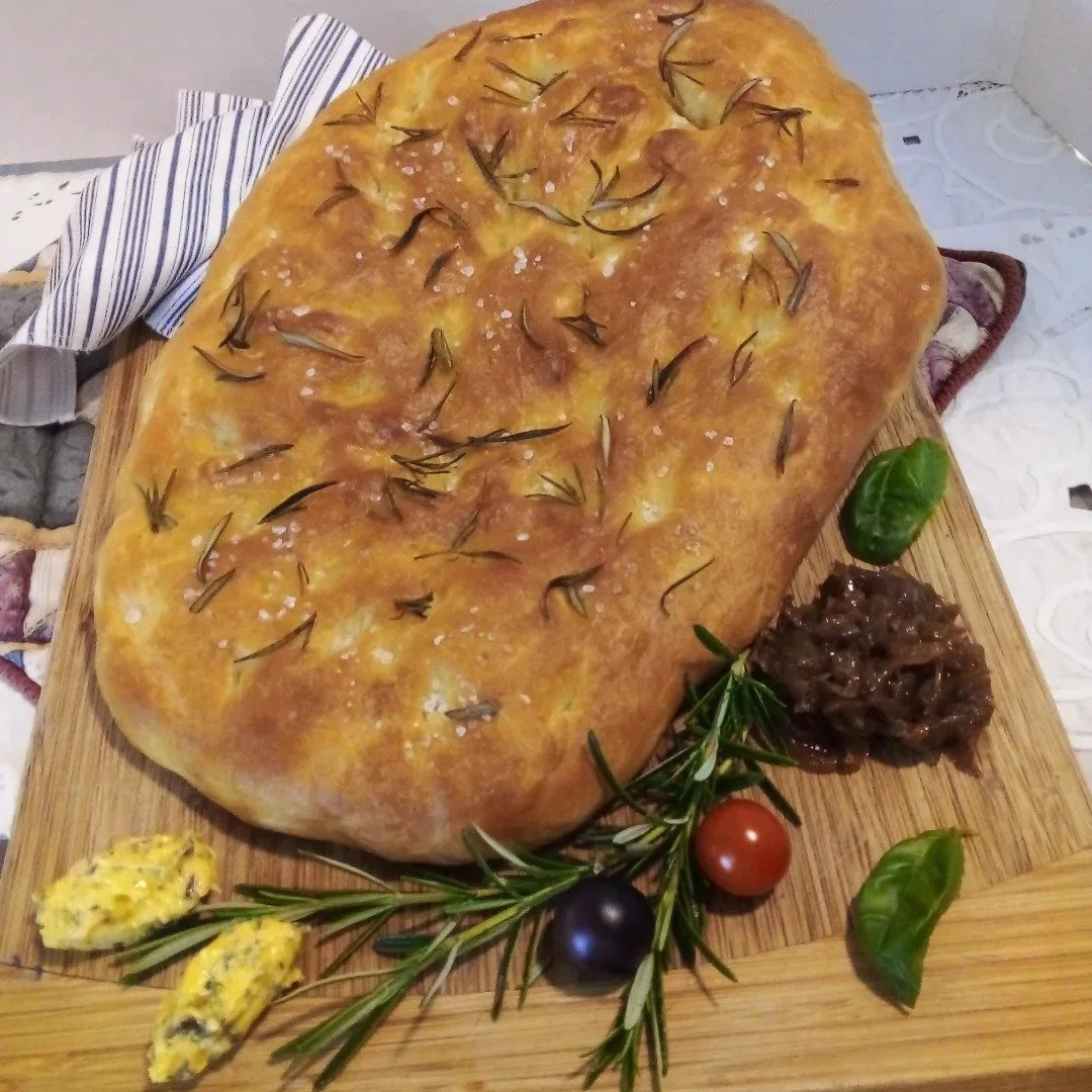 Large round focaccia bread topped with rosemary and coarse salt, served on a wooden board with cherry tomatoes, basil, rosemary sprigs, and a spread of tapenade.