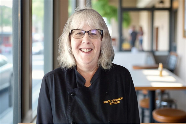 A smiling woman with glasses wearing a chef's coat that says Eden Southern Kitchen, standing inside a restaurant with tables and large windows.