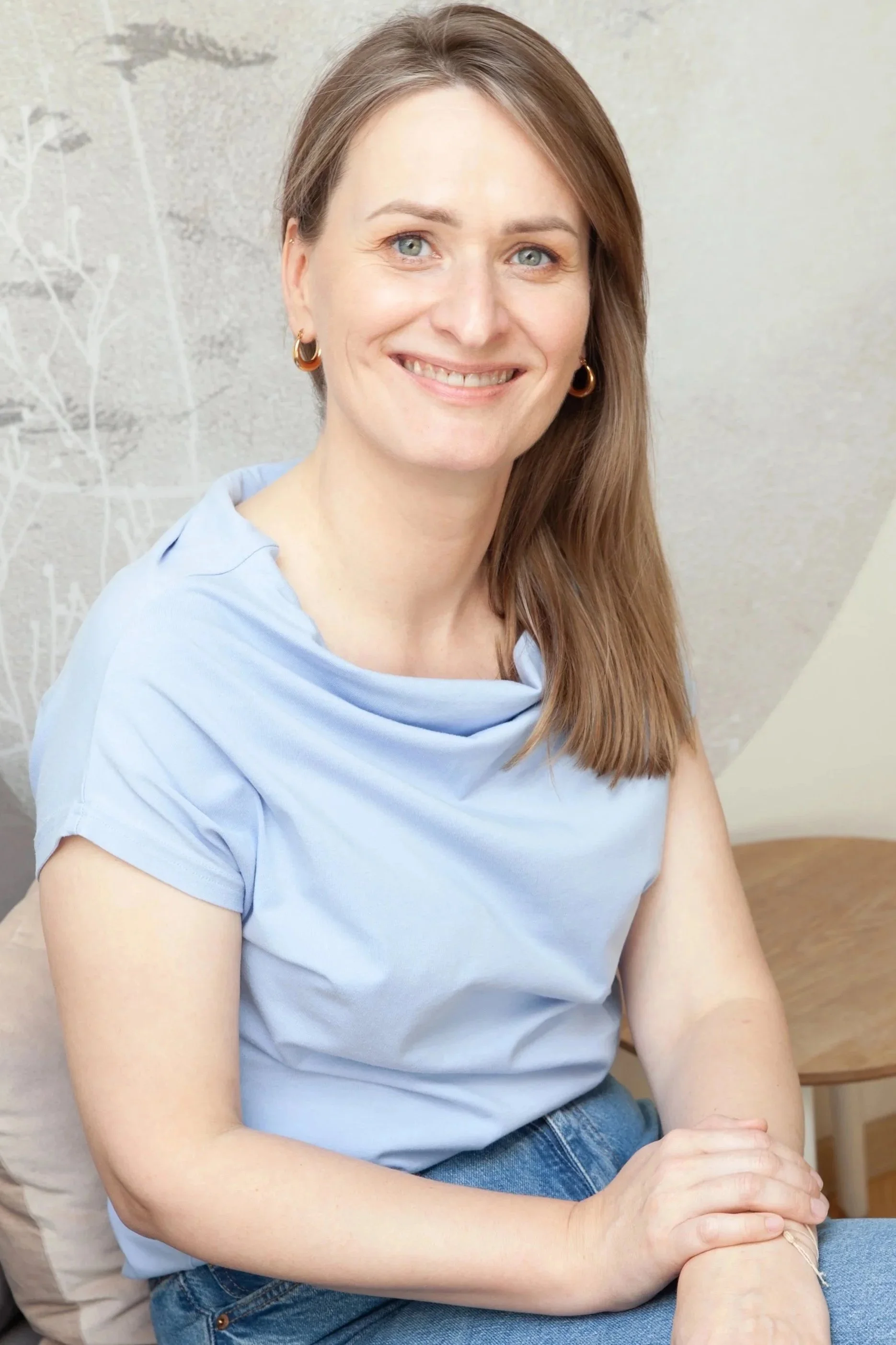 A woman with light skin, blue eyes, and shoulder-length brown hair, smiling and wearing a light blue top and hoop earrings, sitting indoors with a neutral background.