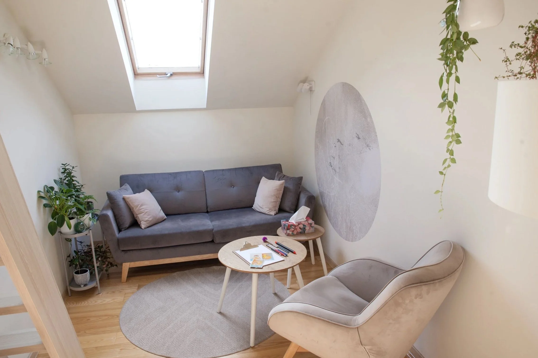 Cozy living room with a gray sofa, beige chair, round coffee table with books and pens, skylight window, and green plants.