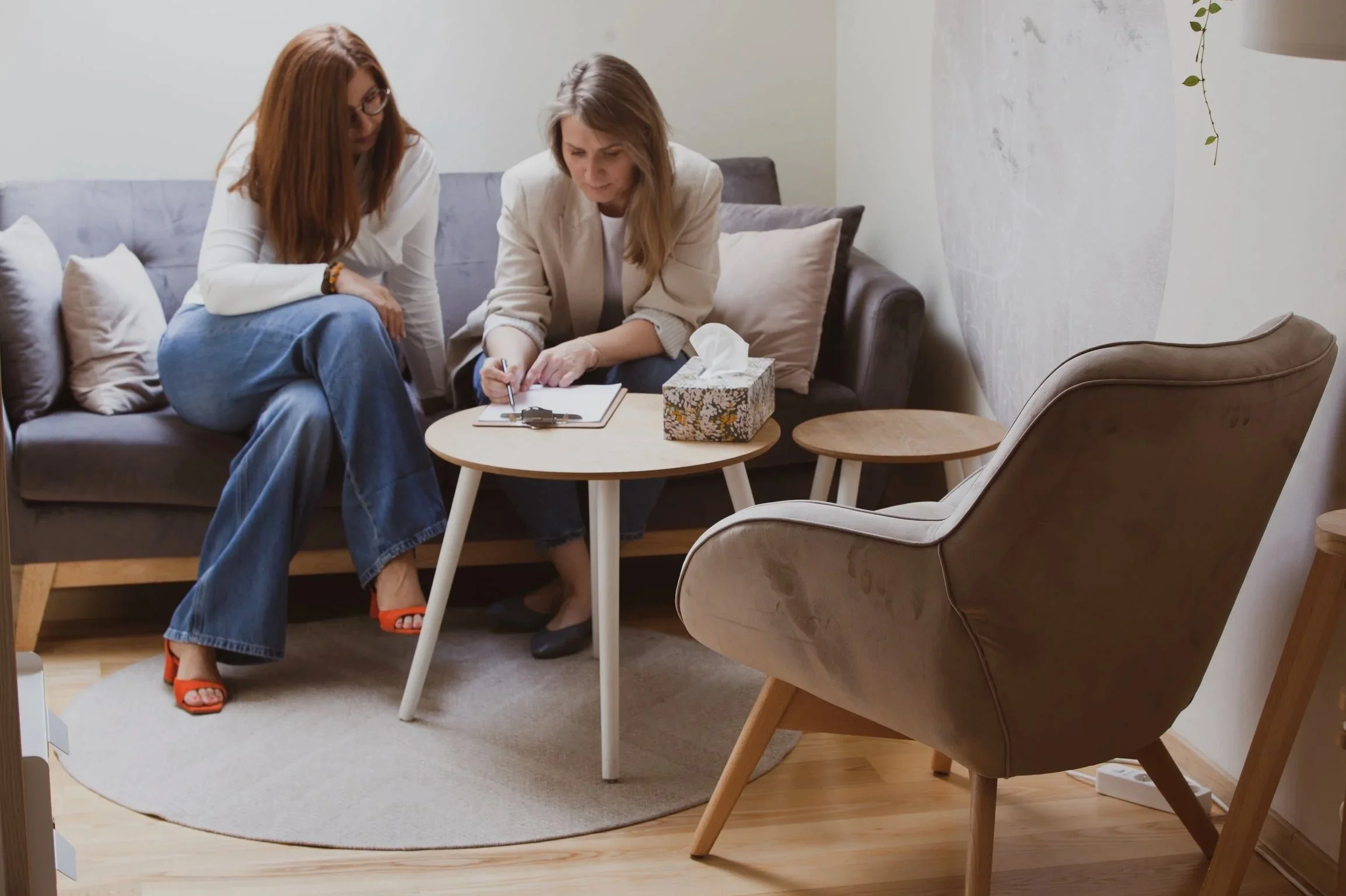 Two women sitting on a gray sofa, with one taking notes on a clipboard, in a cozy living room with a round beige rug and wooden furniture.