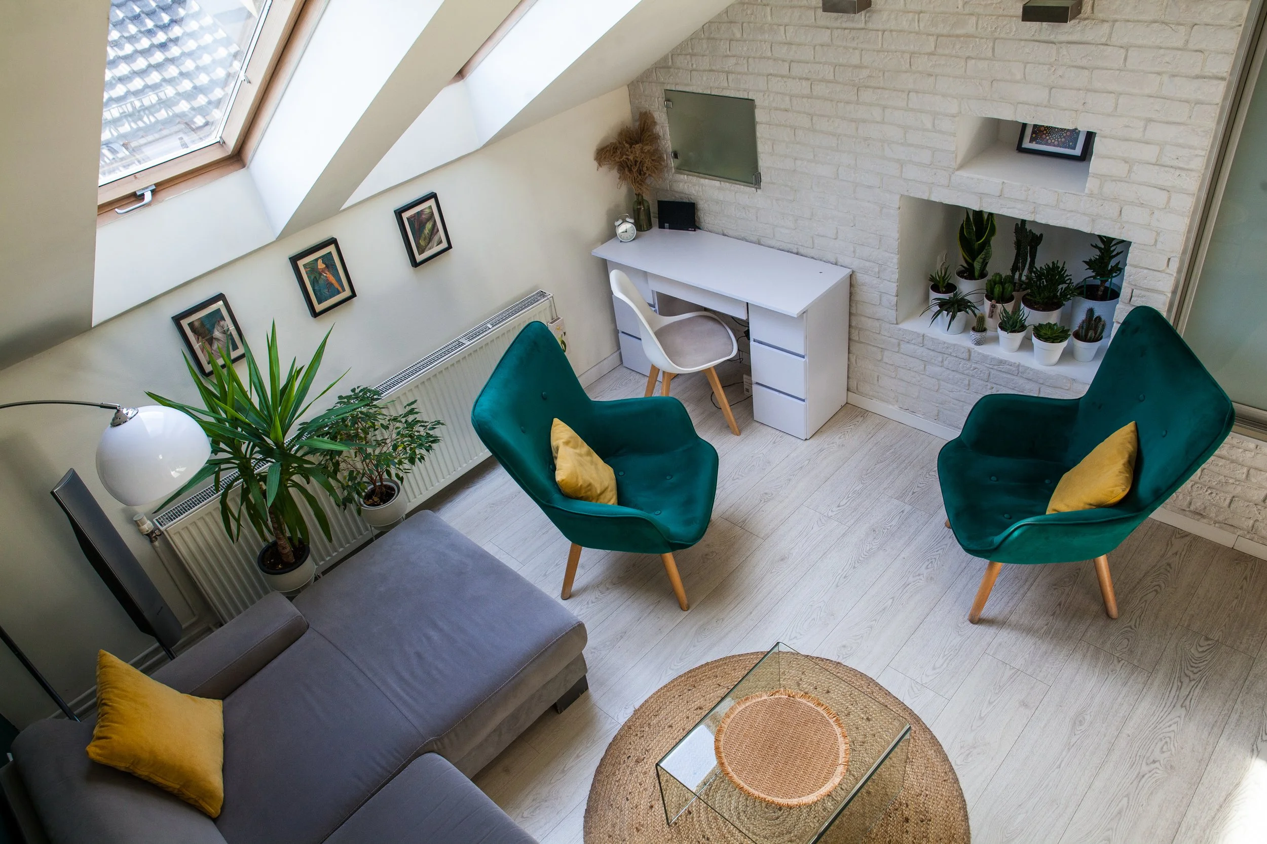 A cozy living room with two teal armchairs with yellow pillows, a gray sofa with a yellow pillow, a round woven rug, a glass coffee table, a white desk with a chair, potted plants, a skylight window, and framed artworks on the wall.