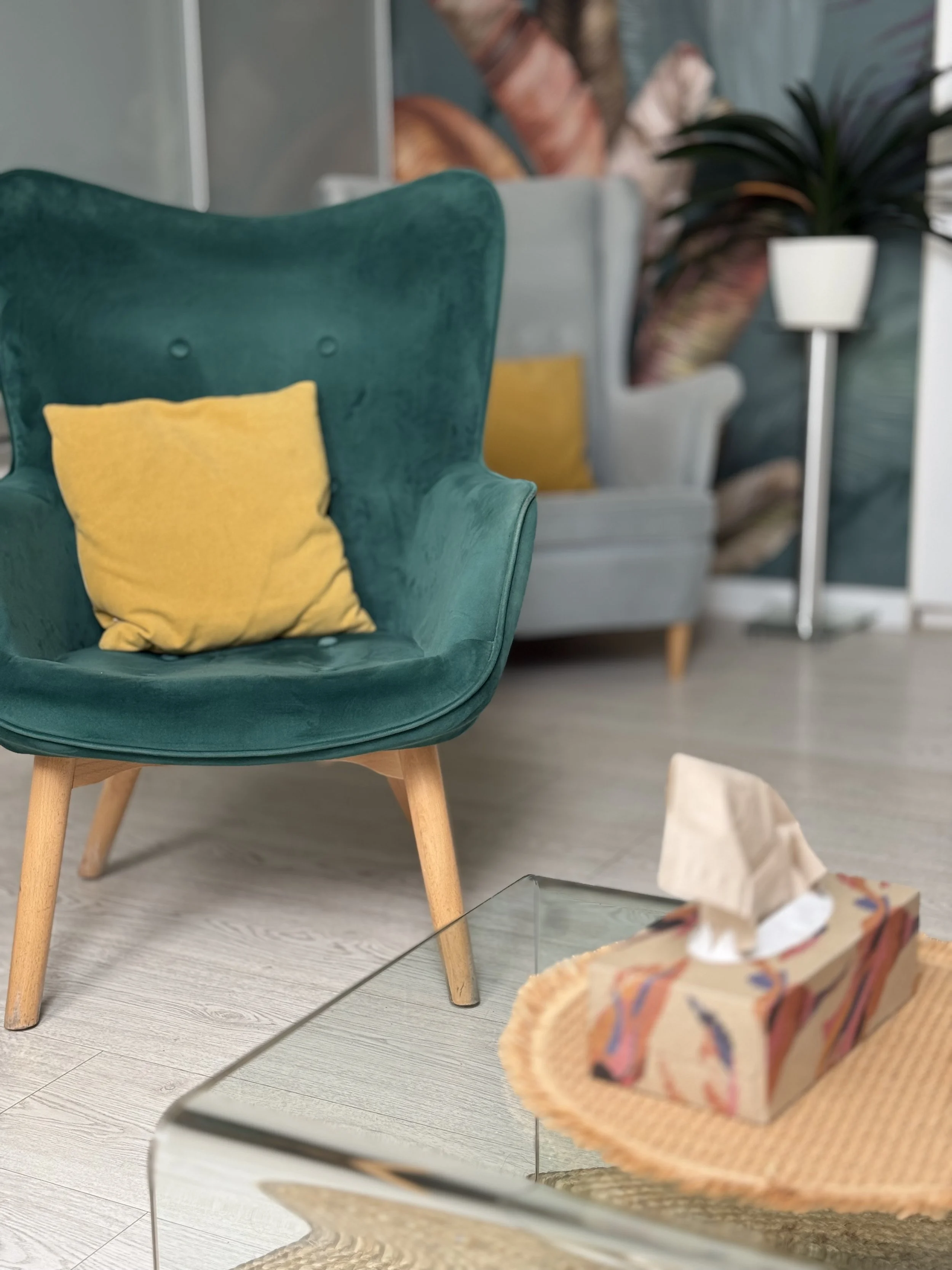 A teal armchair with a yellow cushion, a gray sofa with a yellow pillow in the background, next to a potted plant and a tissue box on a glass table.
