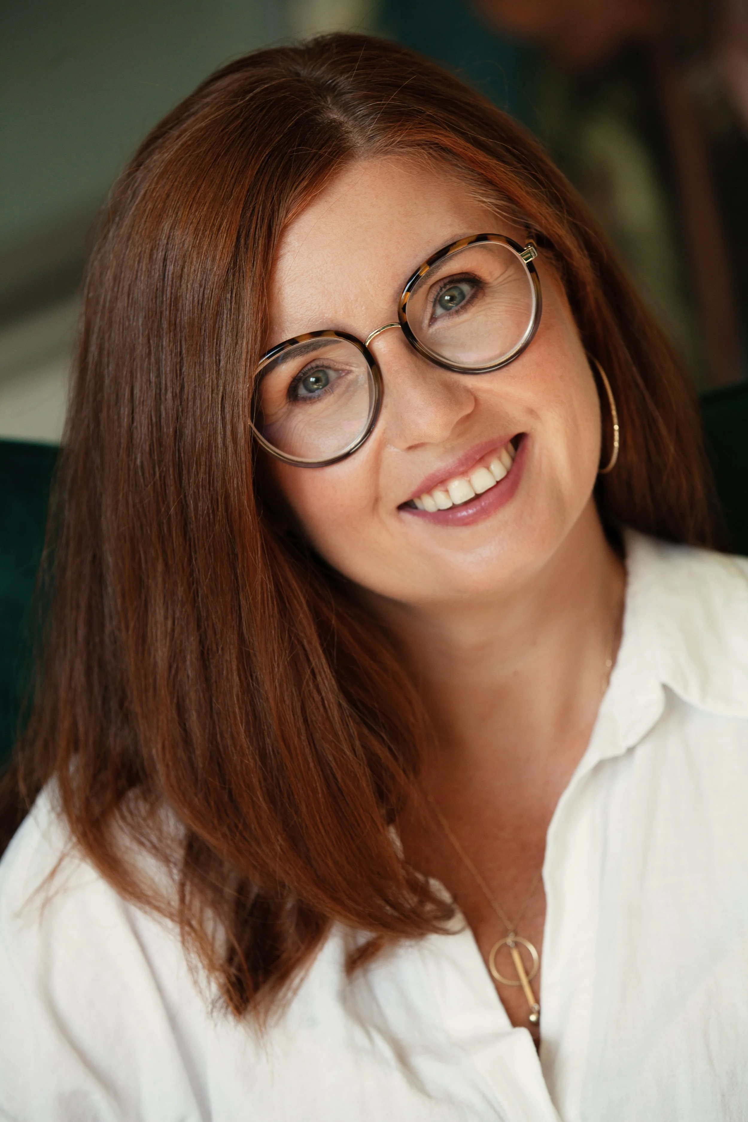 Close-up of a smiling woman with red hair, wearing large round glasses and a white shirt.