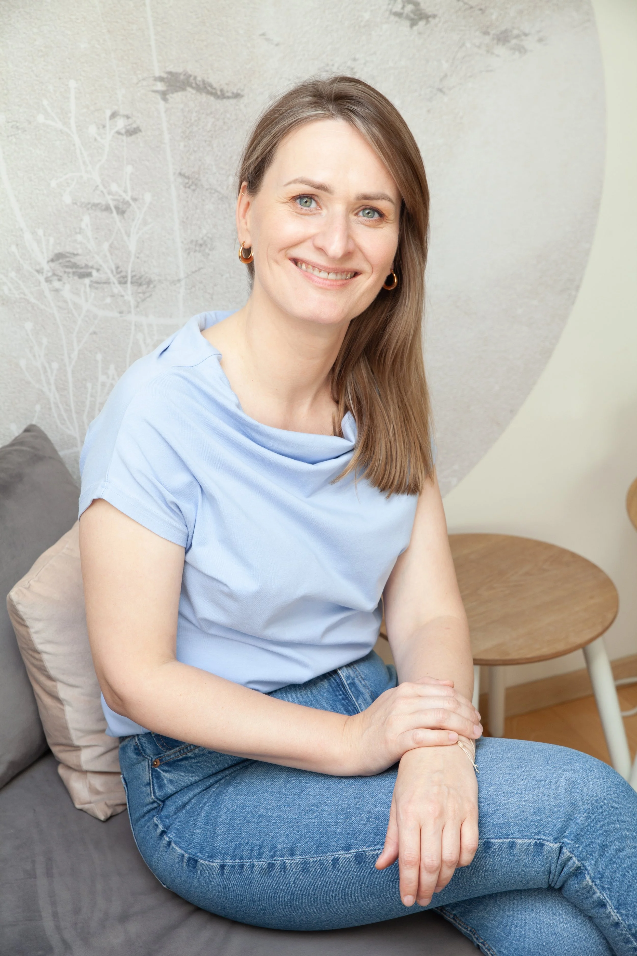 A woman with shoulder-length brown hair and blue eyes is smiling while sitting on a gray couch, wearing a light blue top and jeans in a cozy room with a circular wall decoration and a small round wooden table.