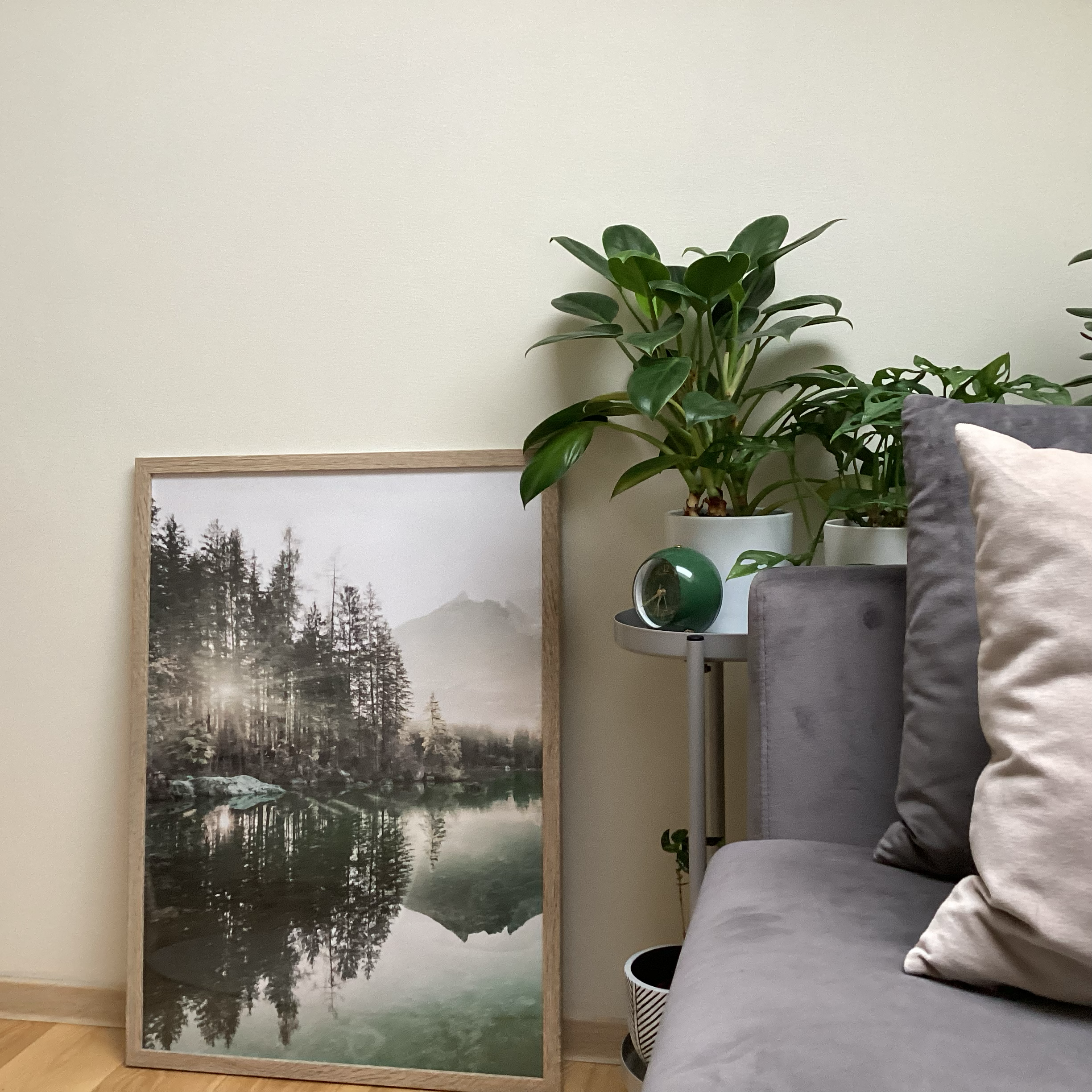 A framed landscape photograph leaning against a wall, showing a lake with trees and mountains, and a reflection of the landscape in the water. Next to it is a small table with potted plants, a green clock, and part of a gray sofa with pillows.