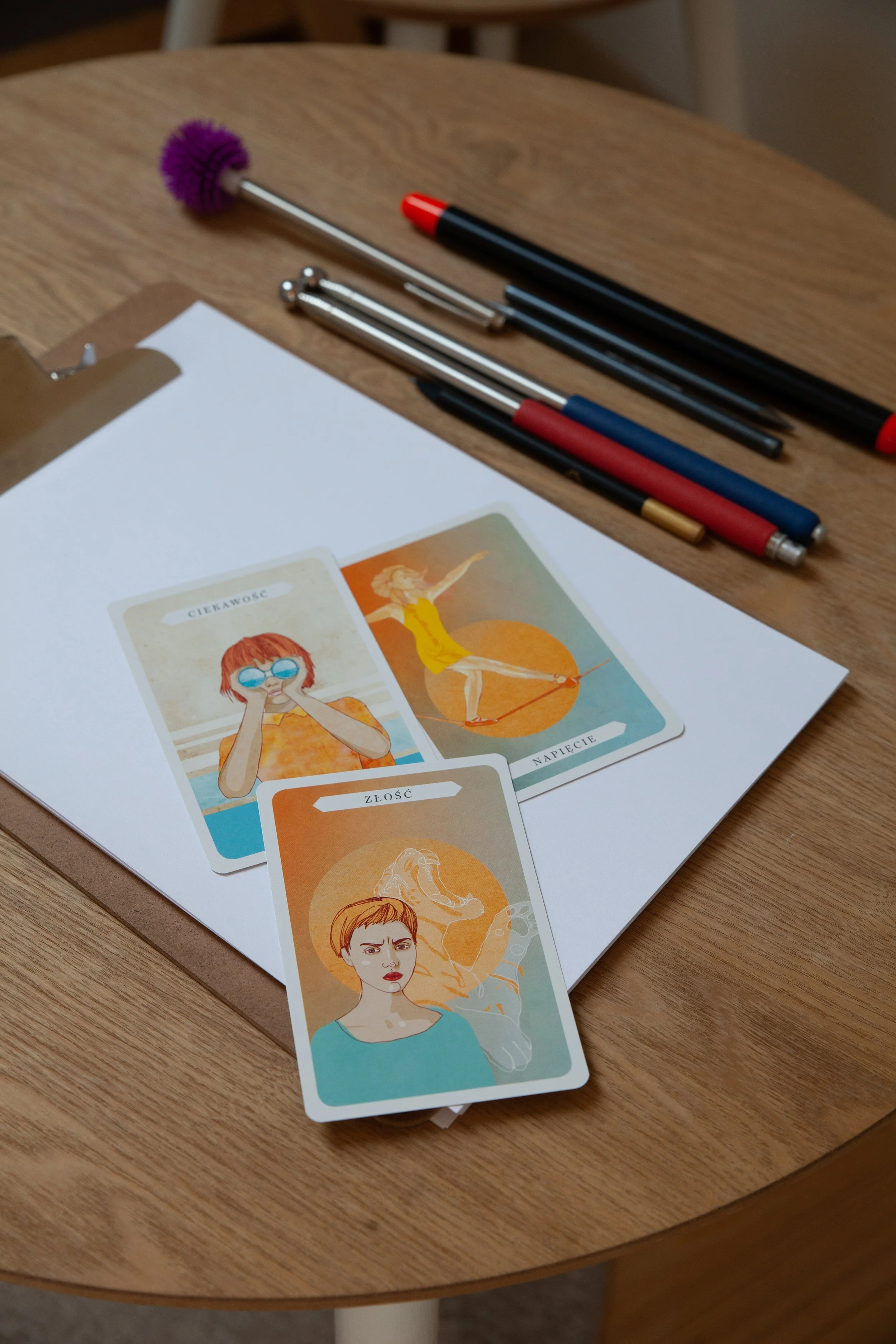 Illustrated cards on a clipboard, depicting emotions such as anger, clarity, and happiness, placed on a wooden table with pens and a purple cleaning brush nearby.