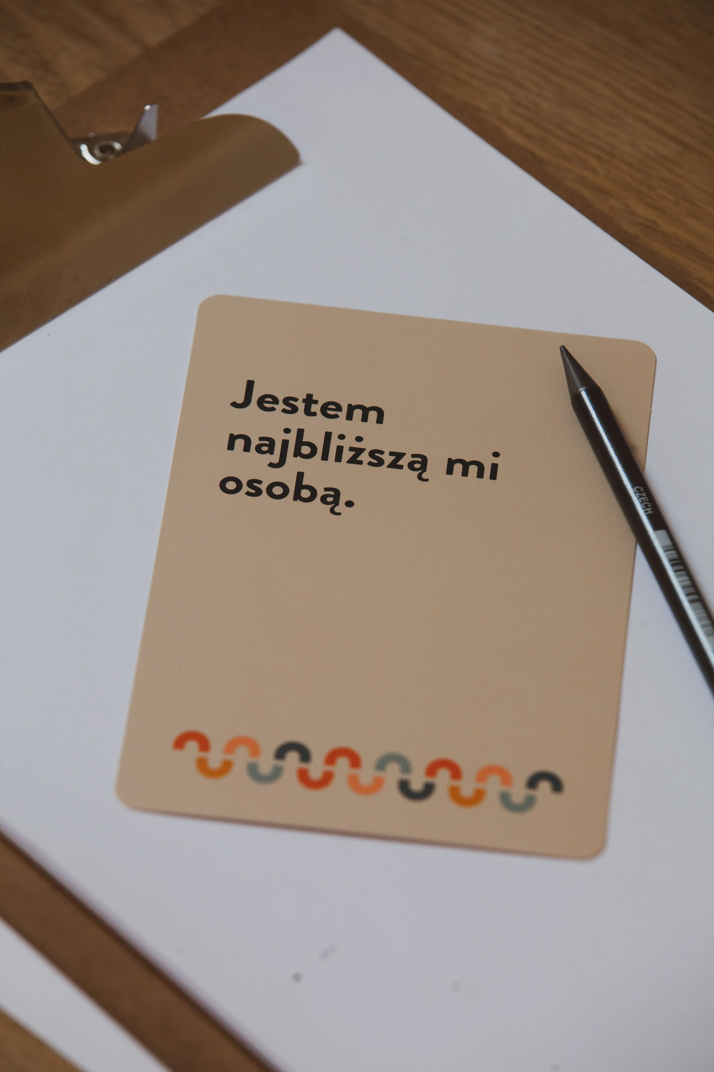 A beige card with Polish text that reads "Jestem najbliższą mi osobą." placed on a white sheet of paper on a wooden desk, with a black pen next to it and a metallic clipboard partially visible at the top left corner.