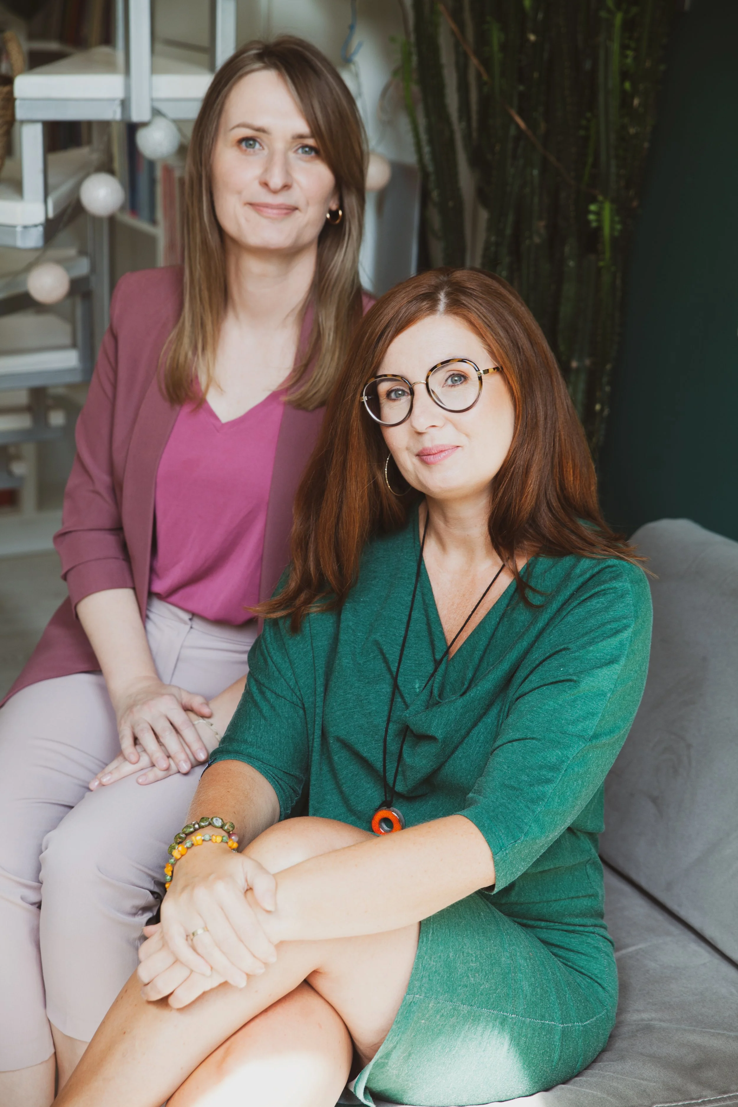 Two women sitting indoors on a sofa, one in a green dress with glasses and shoulder-length red hair, the other in a pink blazer with long brown hair, posing for the camera.