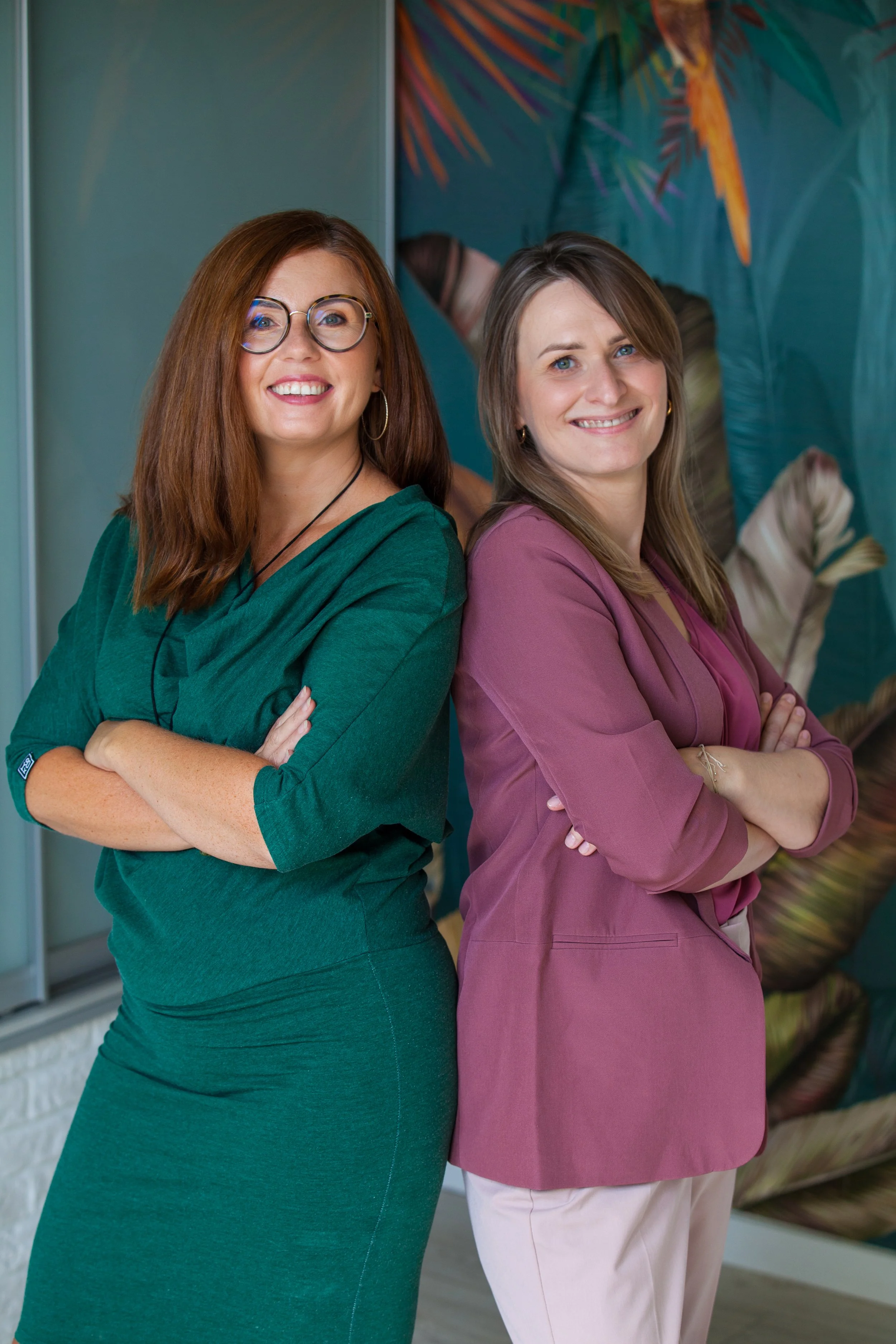 Two women standing back to back with arms crossed, smiling, in a tropical-themed setting.