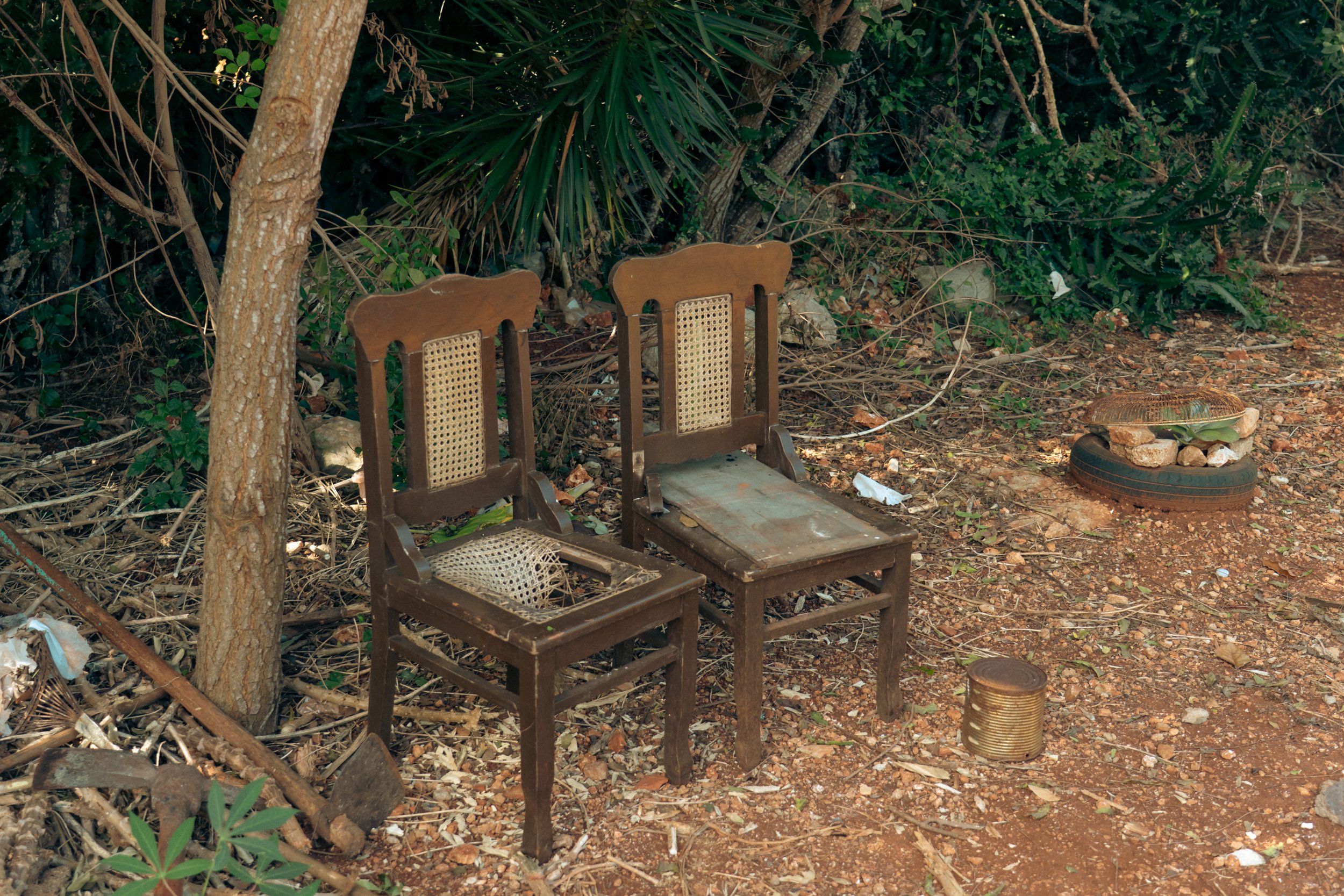 Two old, broken wooden chairs with caned backing, sitting outdoors on a dirt ground with overgrown plants and scattered debris. One chair's seat is missing, and the other has a worn-out wooden plank. To the right, a stone arrangement on a rubber tire