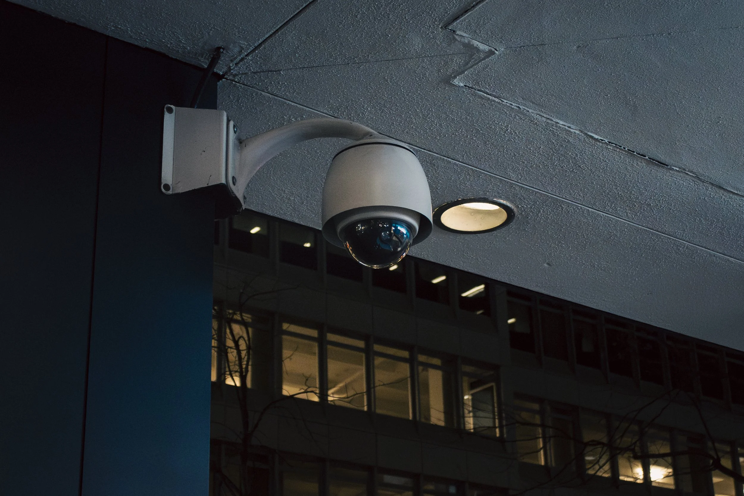 A security camera mounted on the exterior ceiling of a building at night, with a building with lit-up windows in the background.