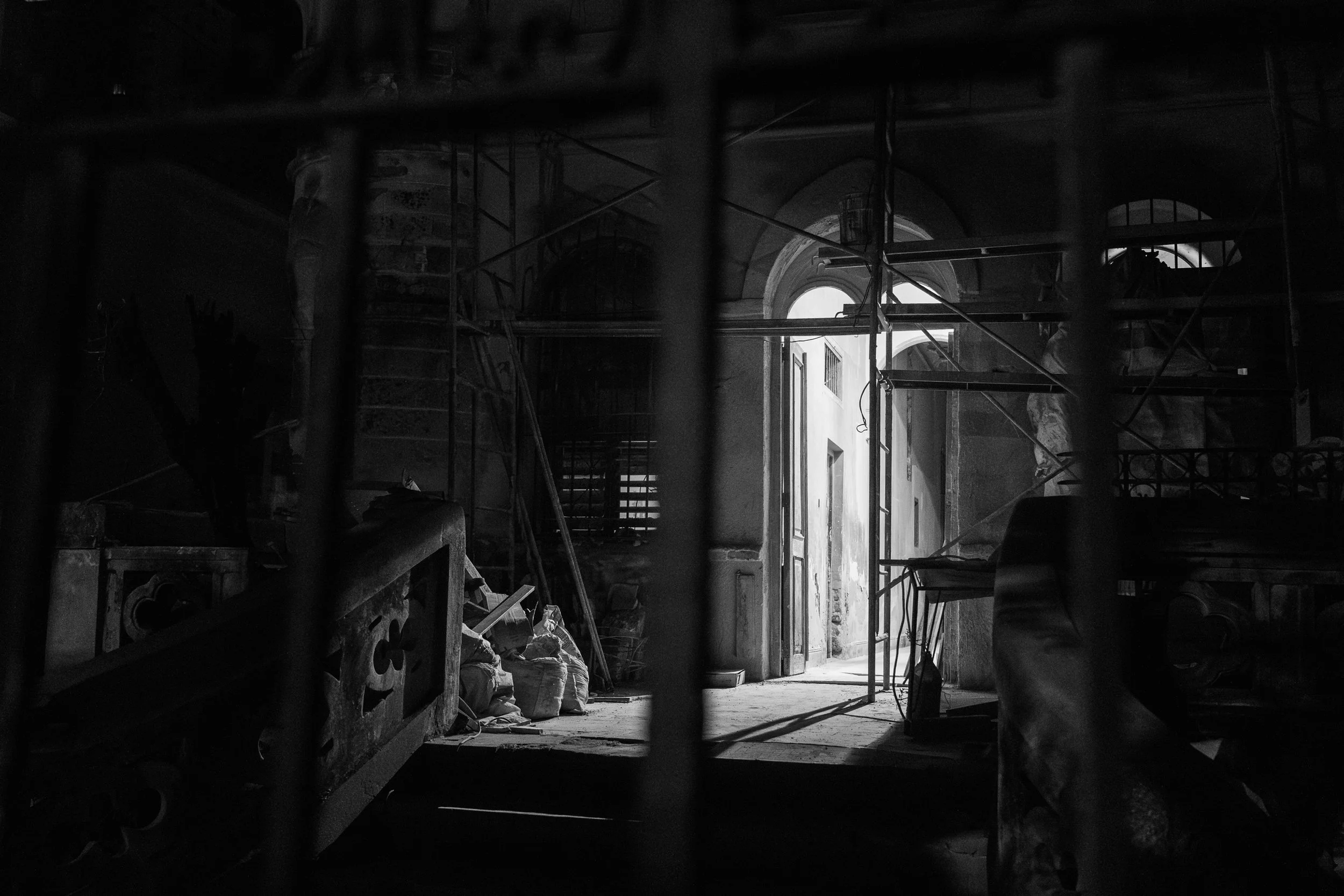 An interior scene with construction scaffolding and building materials, partially obscured by a grid of metal bars. Light comes through an open door and arched windows, illuminating dust and debris on the floor.