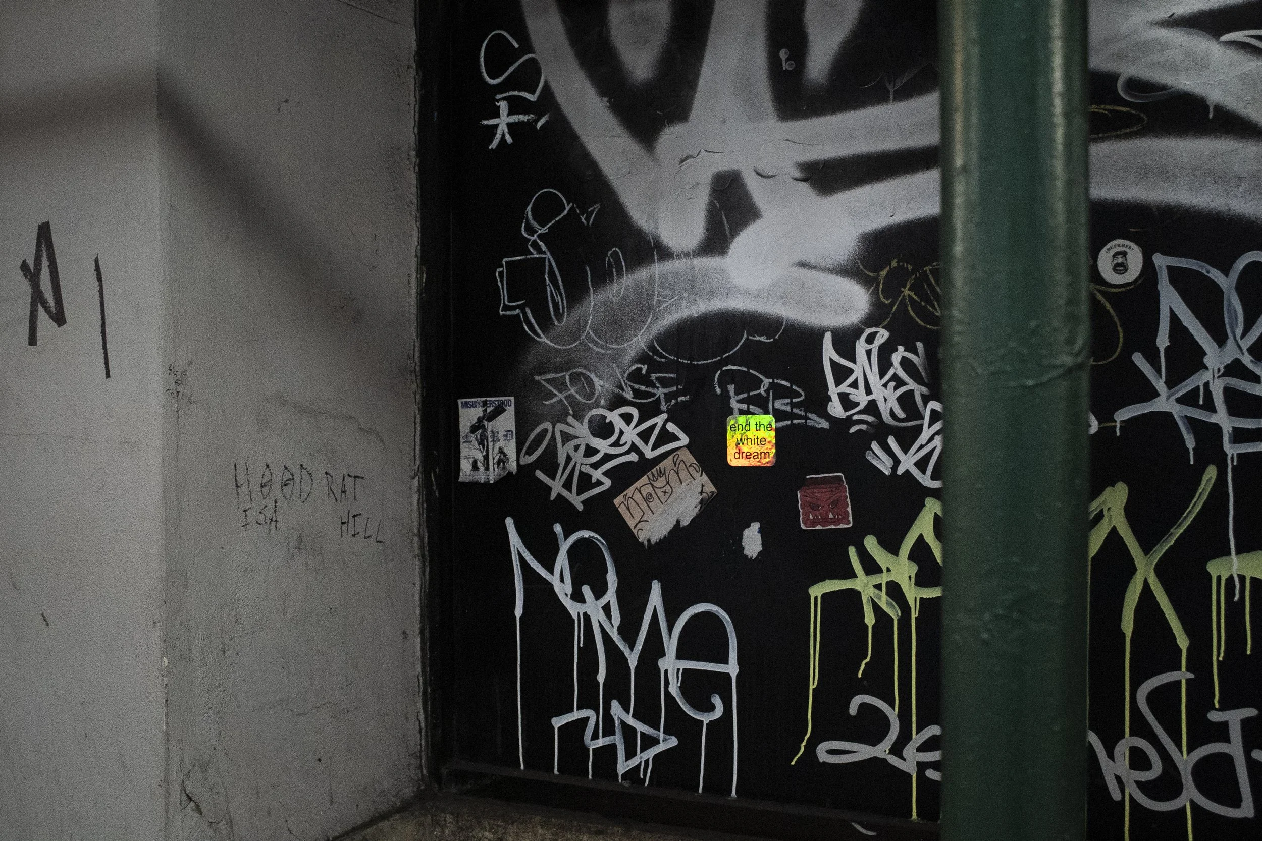 A wall covered with various graffiti, stickers, and street art, including a small sticker with the text 'end the white dream' in yellow and red. There's also a large grey face painted on the wall with dark eyes and nose, partially obscured by a green