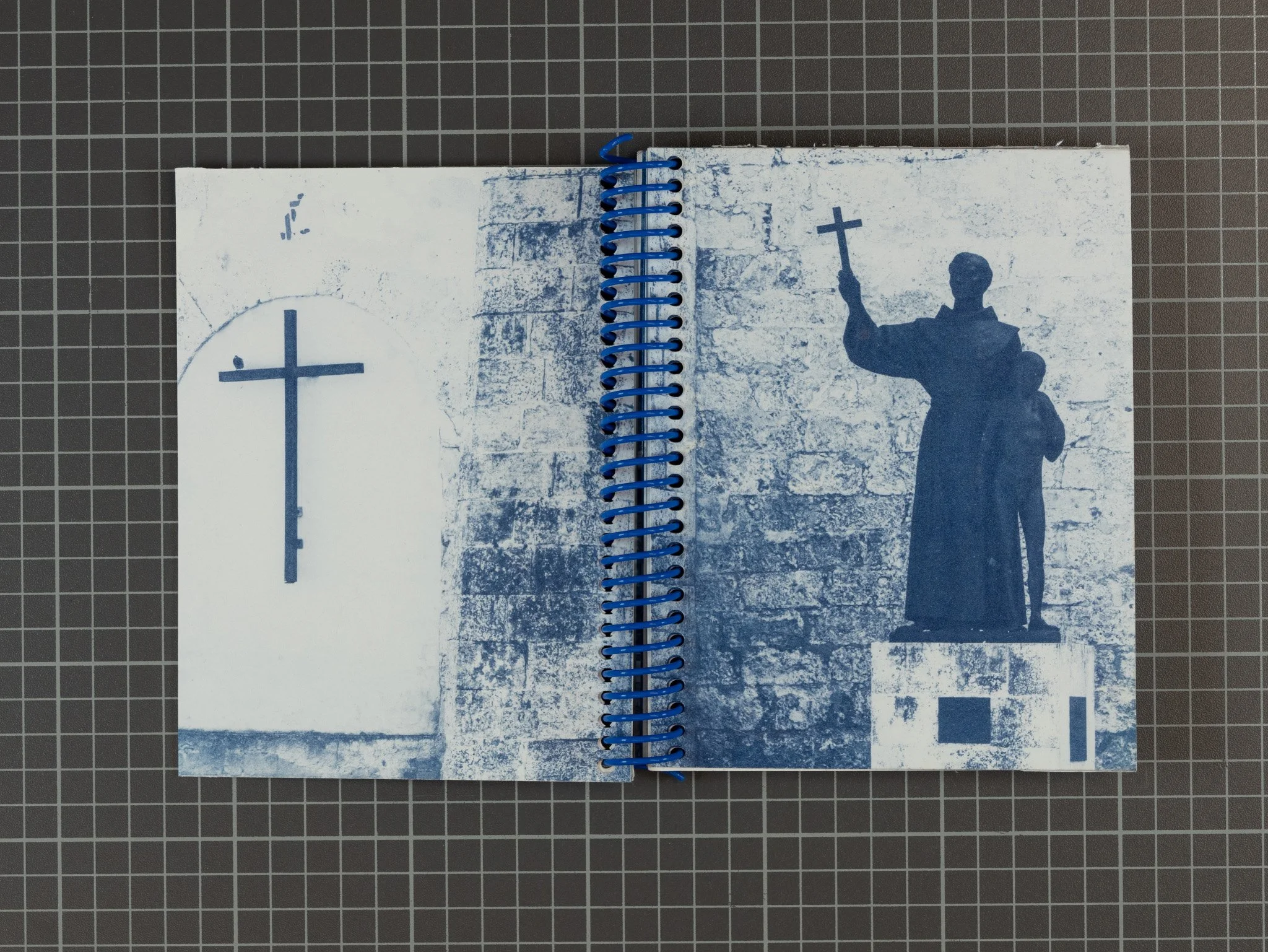 Open spiral-bound cyanotype book with photo of a church cross on the left and a statue of a religious figure holding a cross on the right, set against a textured wall background.