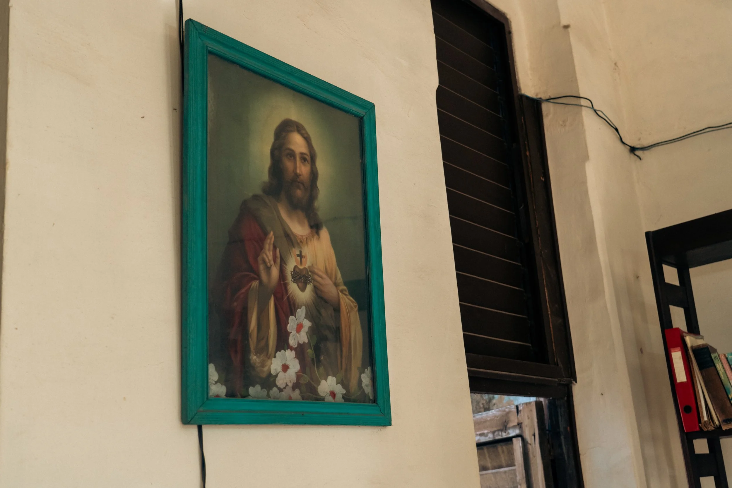 Painting of Jesus Christ with a gesture of blessing, a sacred heart on his chest, surrounded by white flowers, framed in teal, on a white wall.
