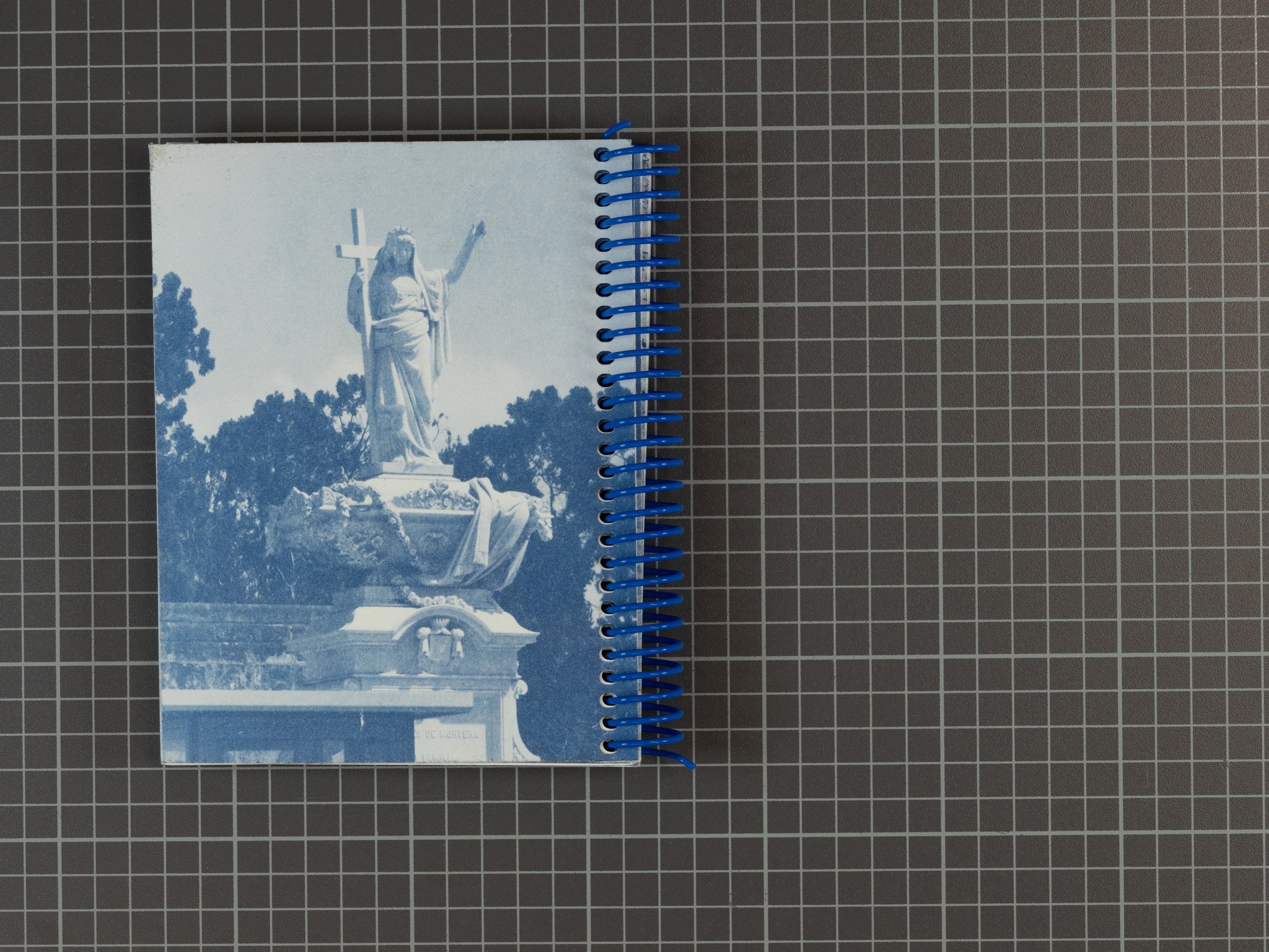 A notebook with a blue spiral binding features a blue-tinted photo of a statue of Jesus Christ holding a cross, situated outdoors with trees around it.