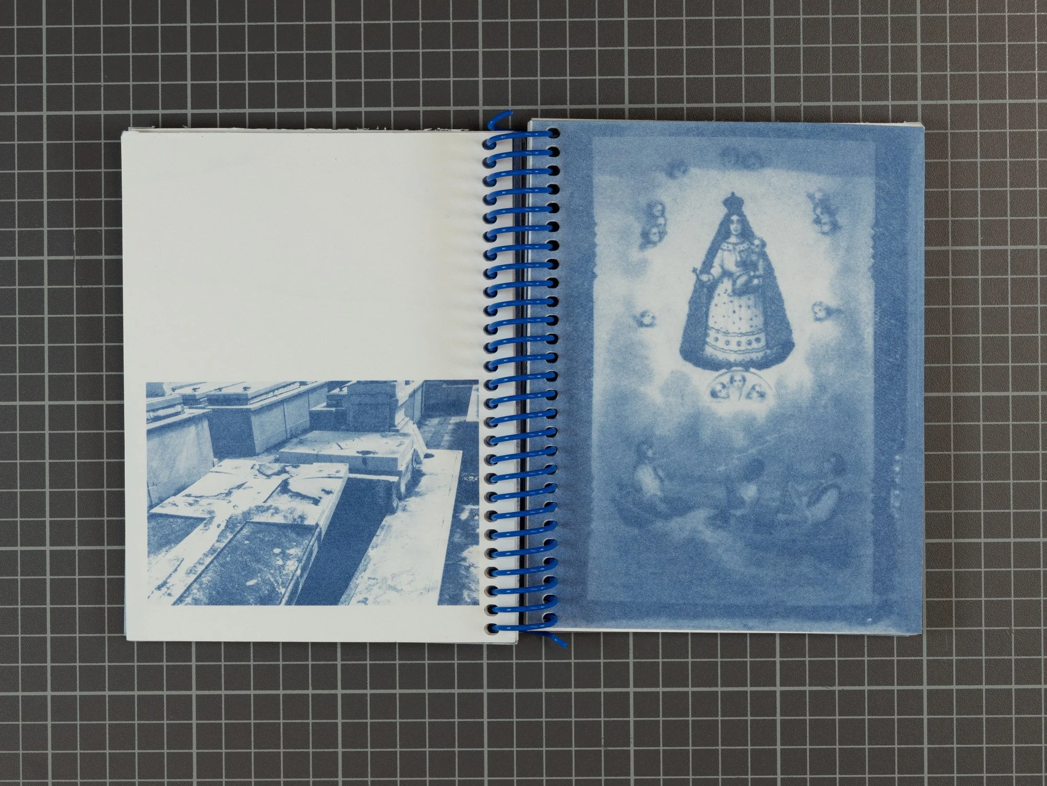Open spiral-bound photo album with two pages; the left page contains a blue-tinted photograph of a table with several items on it, and the right page has a blue-tinted image of a religious icon surrounded by smaller symbols.
