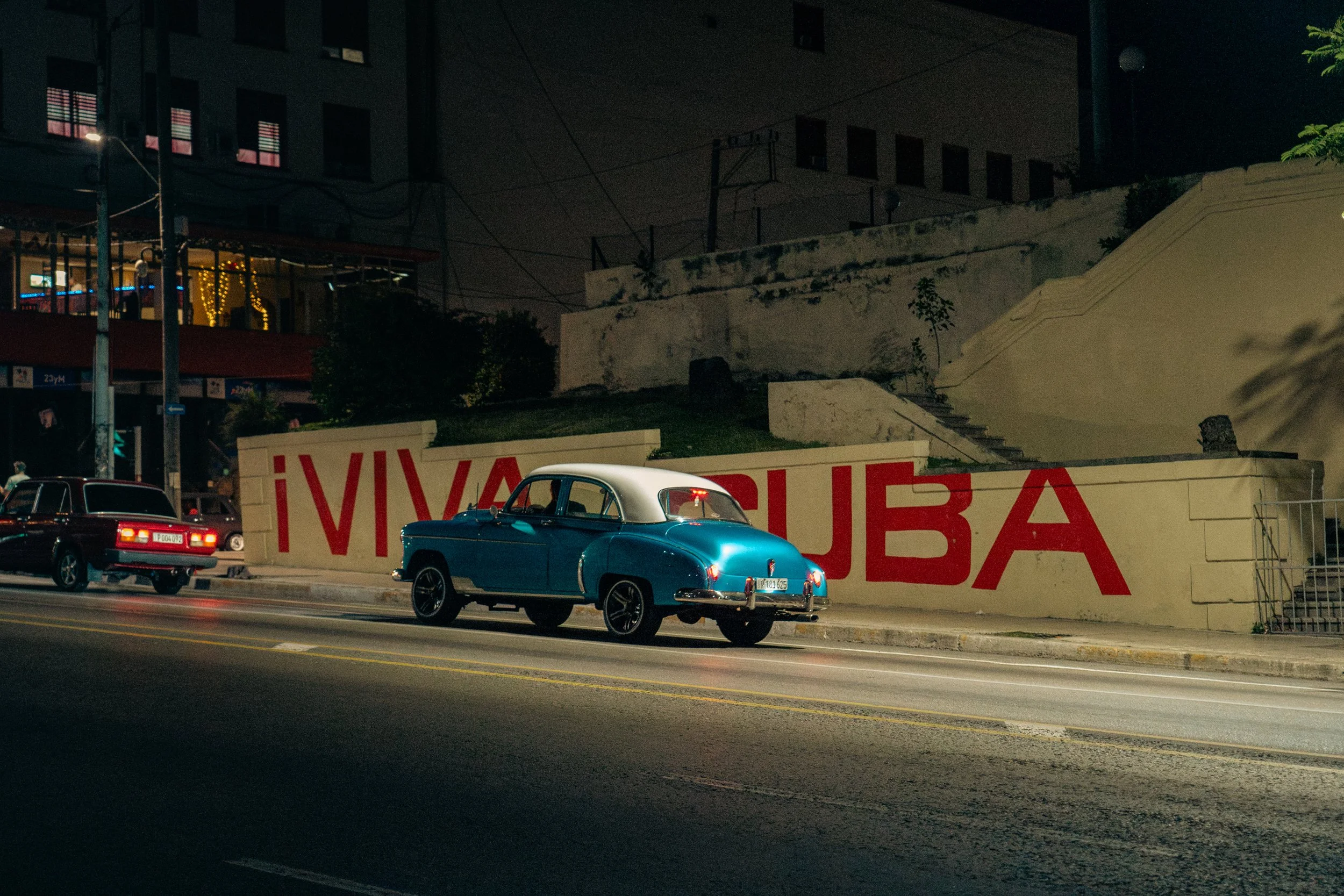 A vintage blue and white car parked along a street at night, behind a wall with red painted letters reading "VIVA CUBA," with another classic car and buildings in the background.