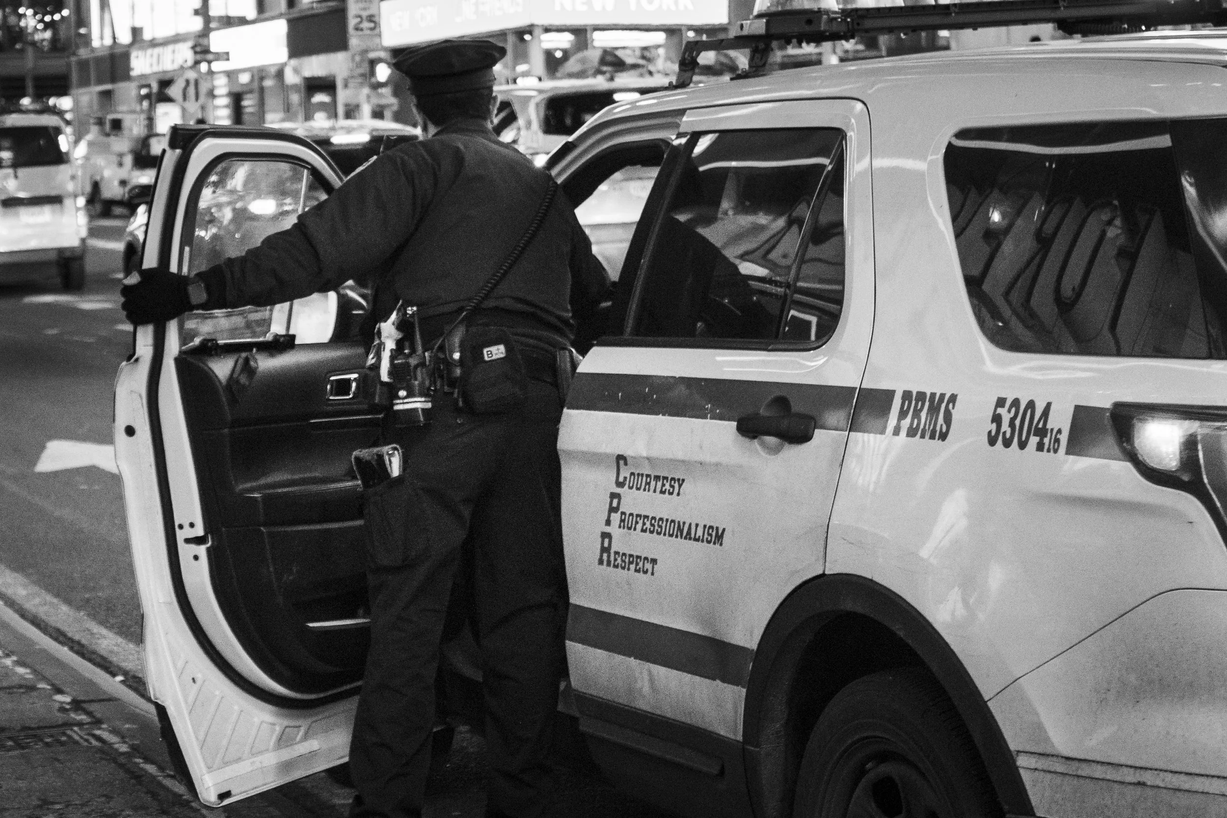 A police officer opening the back door of a patrol car on a city street at night. The car has the words COURTESY, PROFESSIONALISM, RESPECT written on its side.