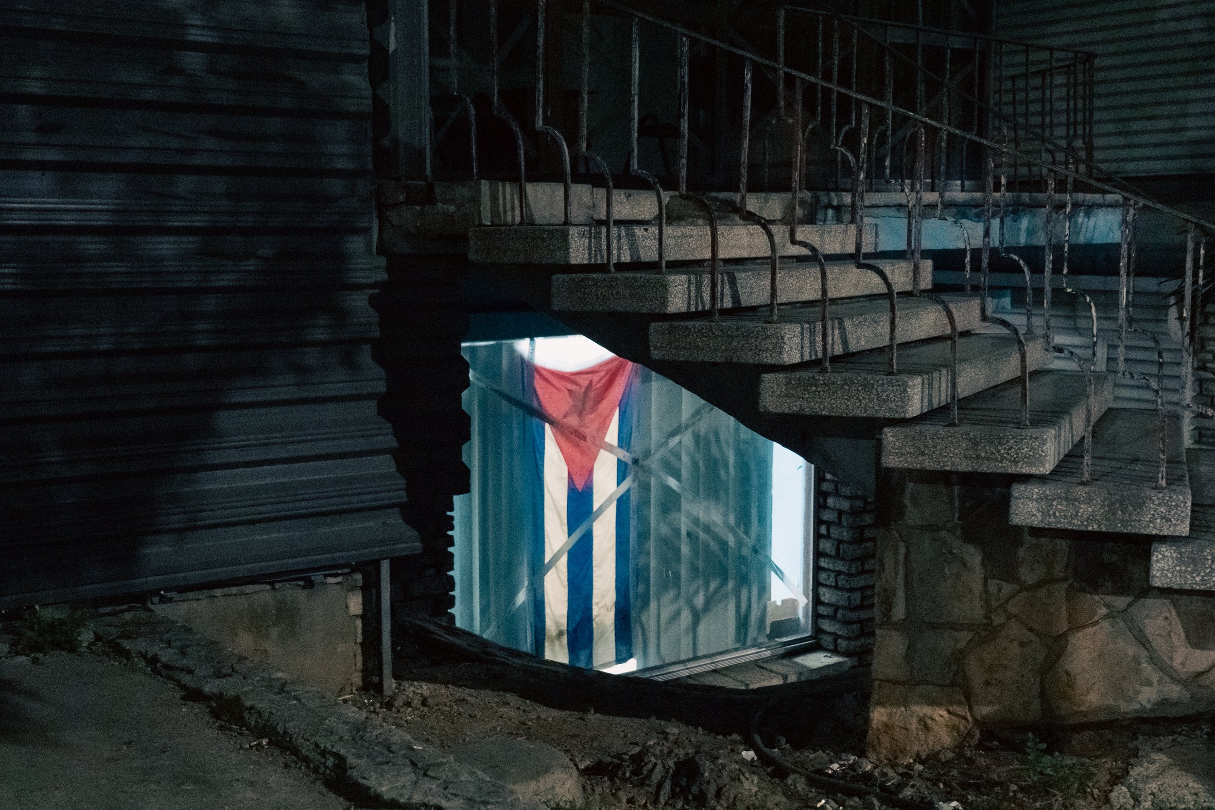A staircase leading down to a basement-like space with a glass door showing the Cuban flag.