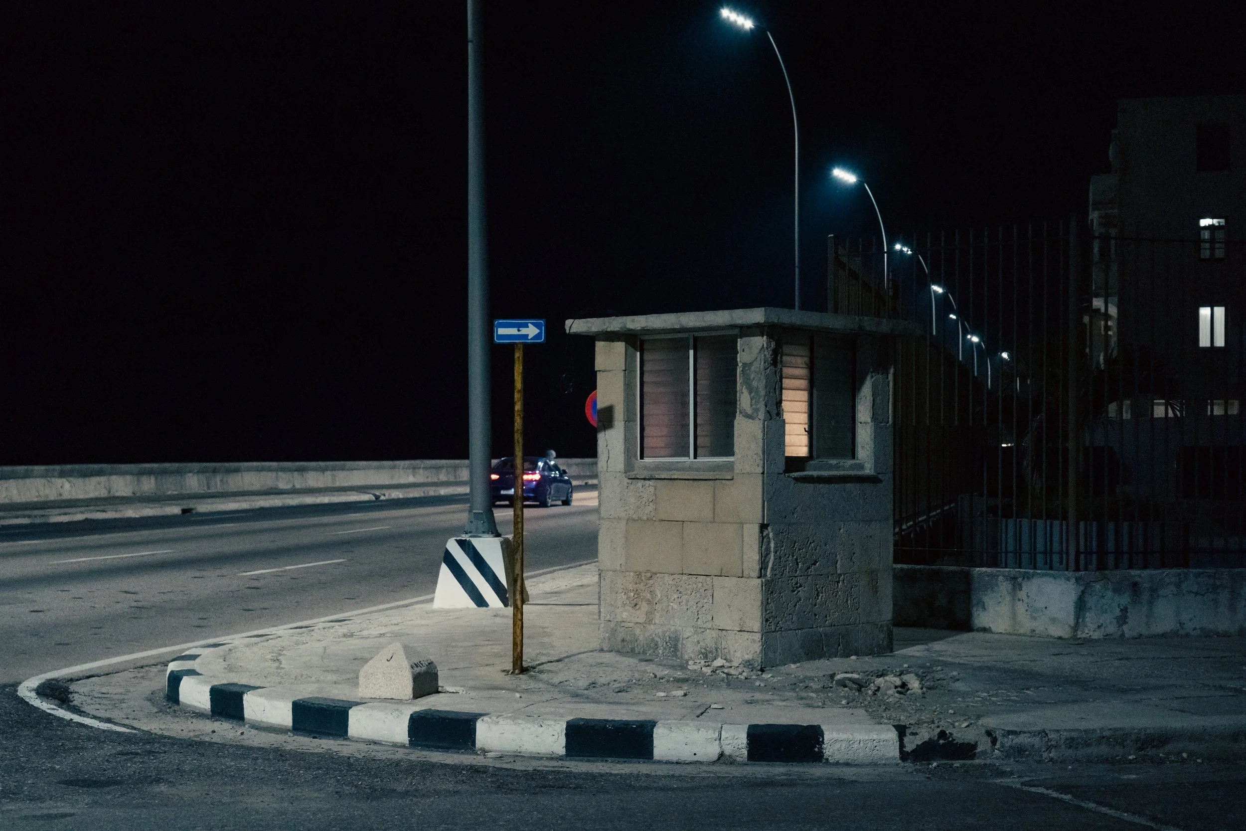 Nighttime view of a small guard booth at the entrance to a gated area, with streetlights illuminating the scene, a car passing by, and a blue street sign with a right arrow.