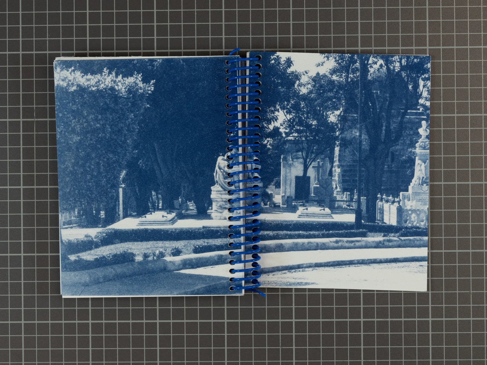 A blue-toned printed photo of a park scene with trees, shrubs, statues, and a platform, presented in an open spiral notebook on a gridded surface.