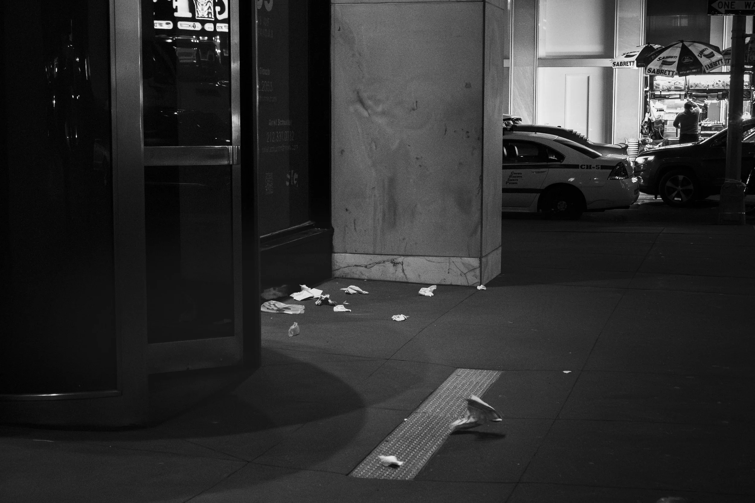 Nighttime city sidewalk with scattered trash including crumpled paper, near a building with large window, some cars and a person with a shopping cart in the background.