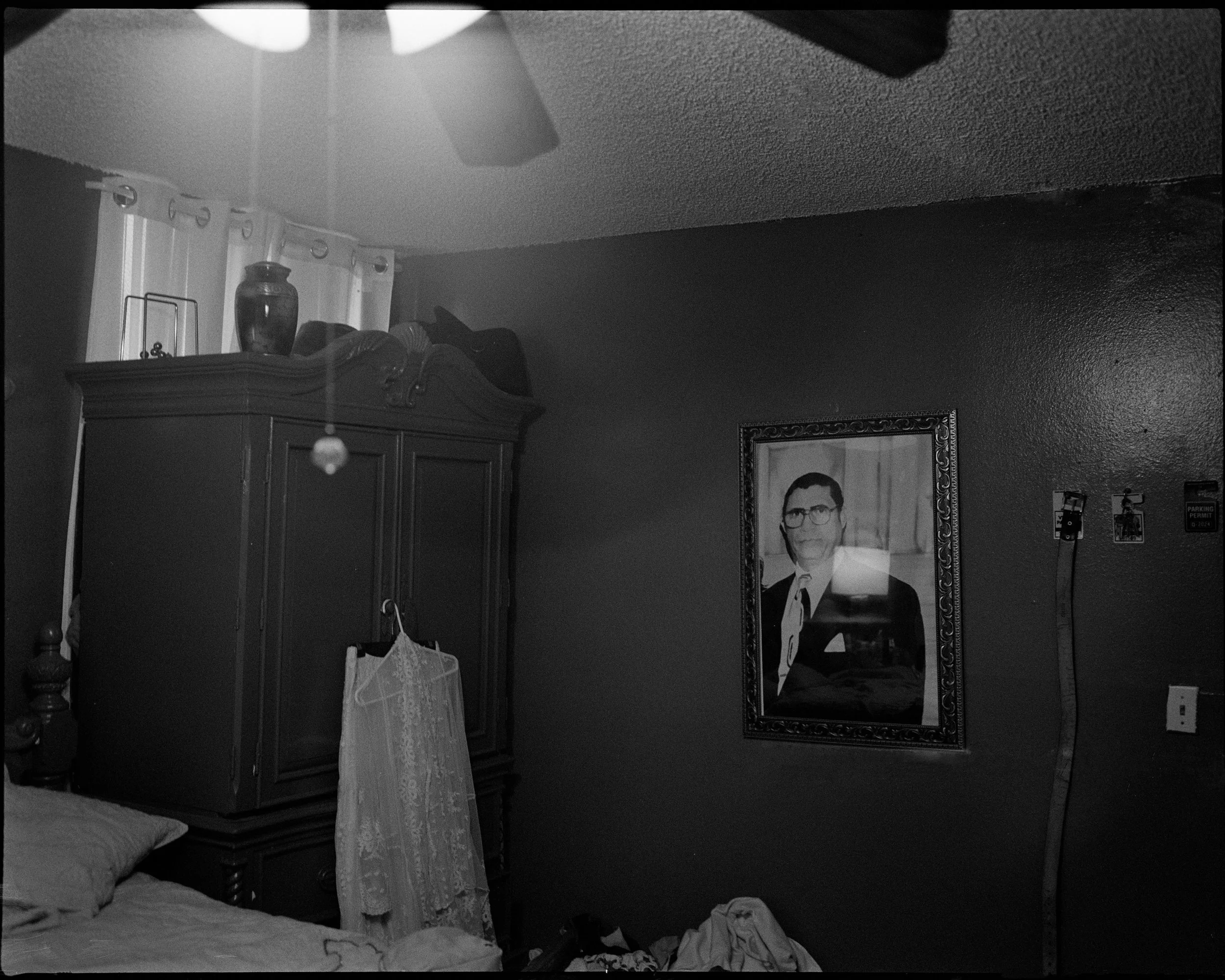 A black and white photo of a bedroom corner showing a bed with pillows, a dark wooden wardrobe with a lace dress hanging from it and a framed portrait of recently departed grandfather.