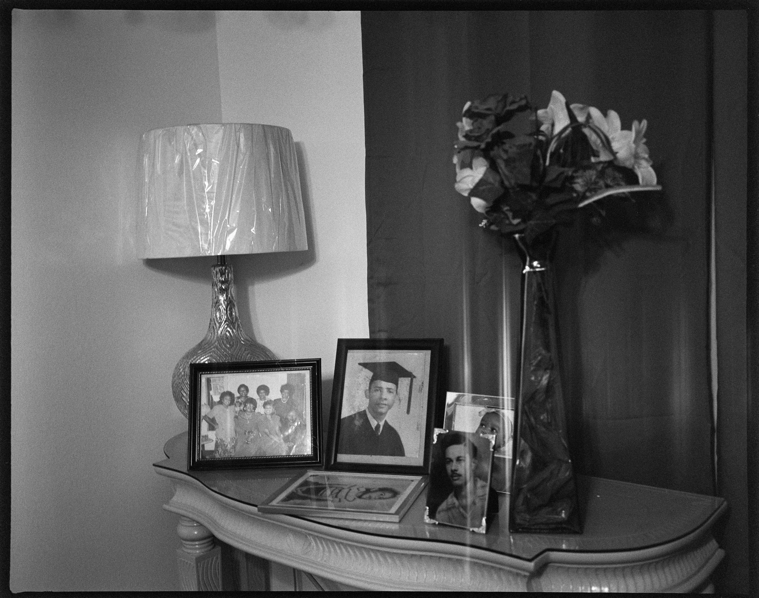 A table with old framed family photographs, a vase with flowers, and a lamp in a living room.