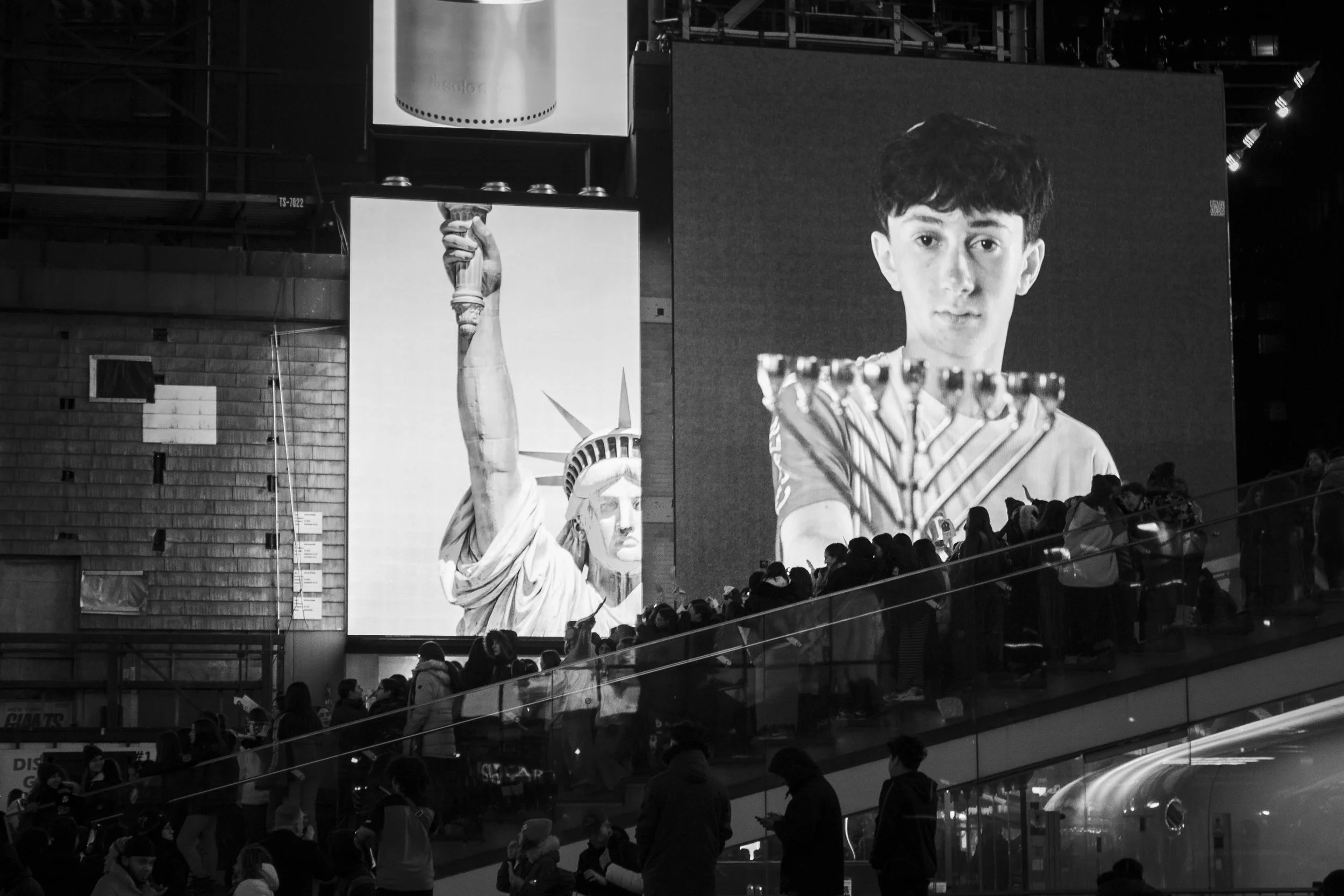 A large digital display showing an image of the Statue of Liberty and a boy holding a menorah with nine candles. People are walking and gathering in a modern indoor space with a staircase in the foreground.