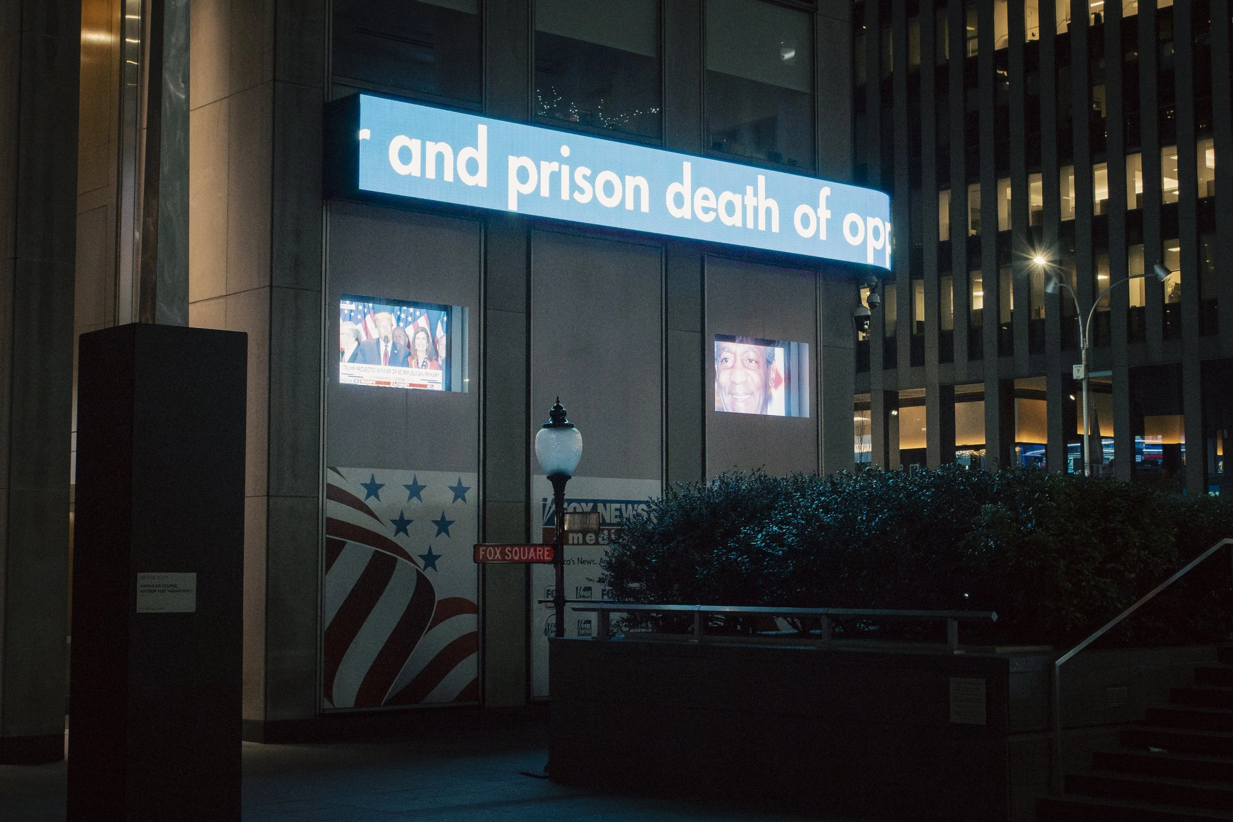 Nighttime scene of an urban building with large illuminated sign reading 'and prison death of op'. Two screens display news footage and a face, along with a Bush logo and American flag-themed graphic, at Fox Square in New York City.