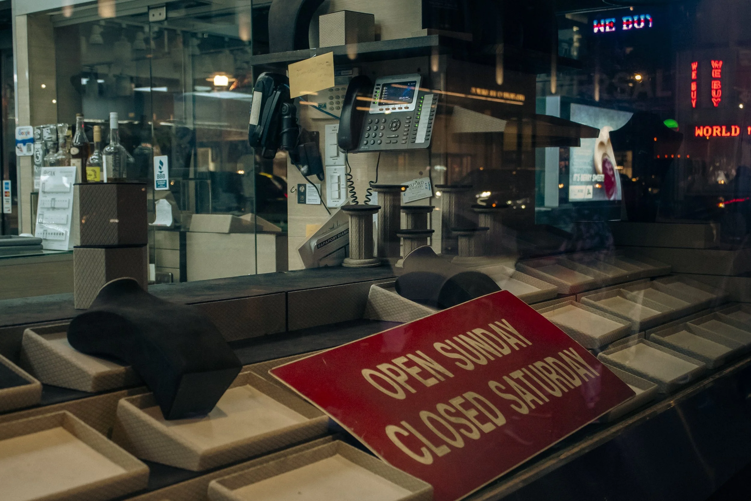 View of a closed jewelry store display window with an 'Open Sunday, Closed Saturday' sign, empty jewelry trays inside, and reflections of city lights outside.