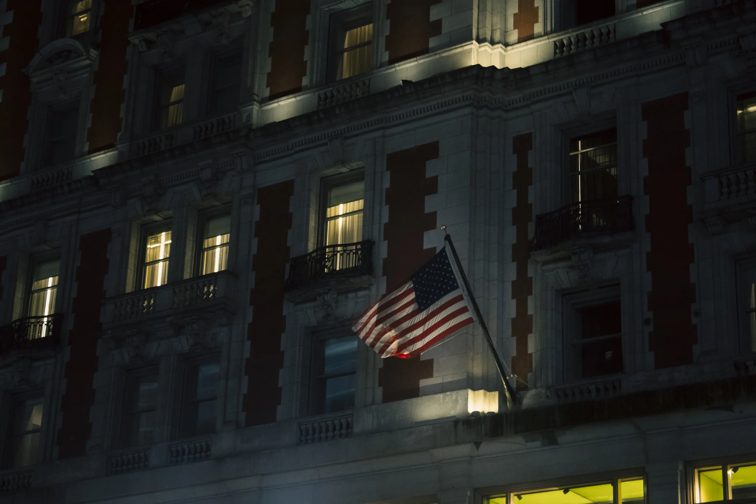 Nighttime view of a building with lit windows, featuring an American flag hanging from a pole attached to the building.