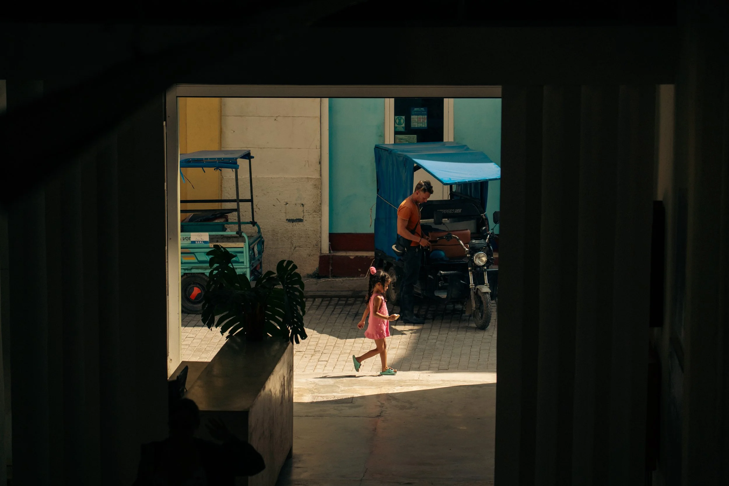A young girl in a pink dress walking outside on a sunny day, with a man and a tricycle in the background, partially shaded by an overpass or structure.